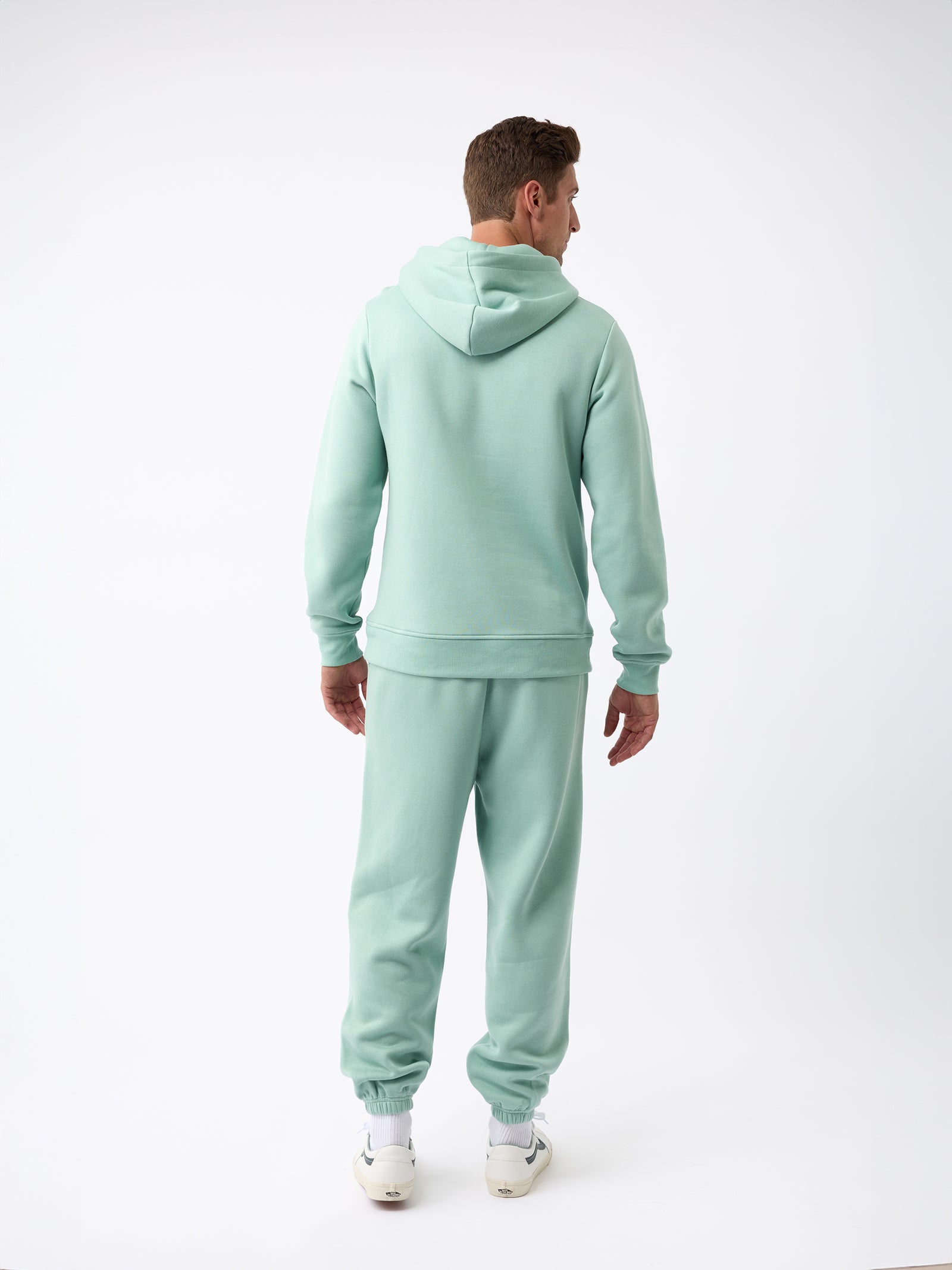 A person dressed in a Men's CityScape Sweatpant from Cozy Earth is facing away from the camera. They are standing against a plain white background, showcasing their light green hoodie and matching sweatpants, along with visible white sneakers.