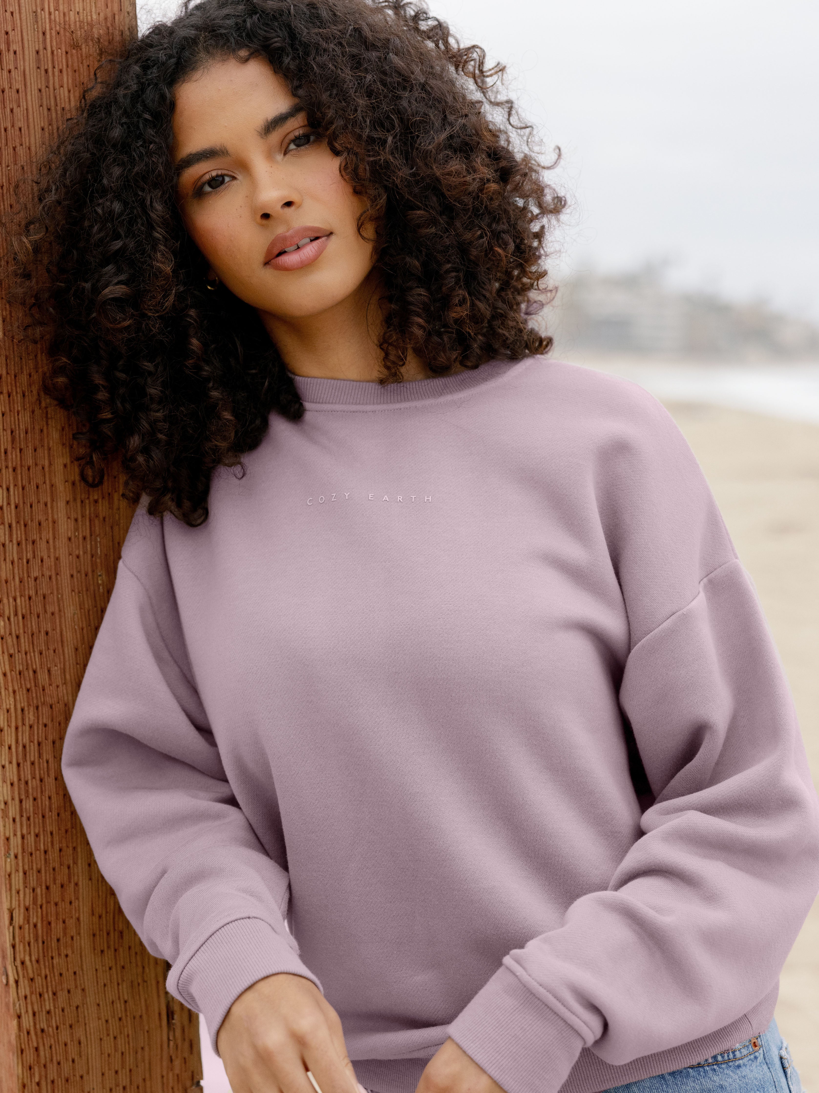 Dusty Orchid CityScape Pullover Crew. The Pullover is being worn by a female model. Accompanying city scape clothing is being worn to complete the look of the outfit. The photo was taken with a sandy beach background. |Color:Dusty Orchid