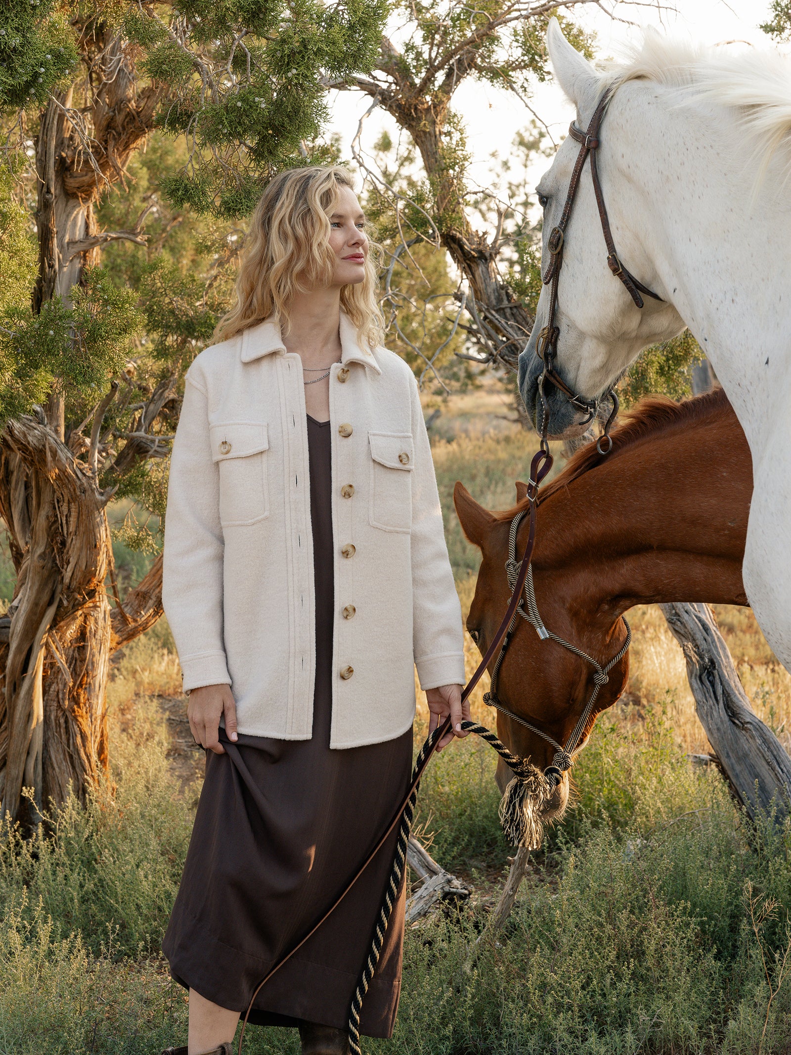 A person with wavy blonde hair stands outdoors wearing a Cozy Earth's Women's Boucle Shacket in beige over a brown dress, holding the reins of two horses. The background features trees and grass, suggesting a natural setting.