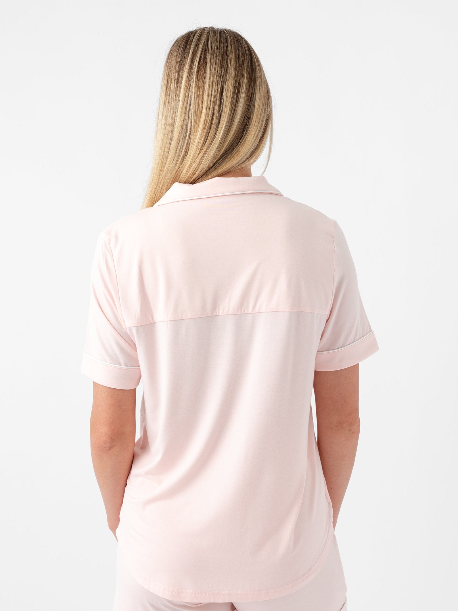 A person with long blonde hair is shown from the back, wearing a light pink Women's Short Sleeve Bamboo Pajama Top in Stretch-Knit by Cozy Earth, along with matching shorts. The background is plain white.