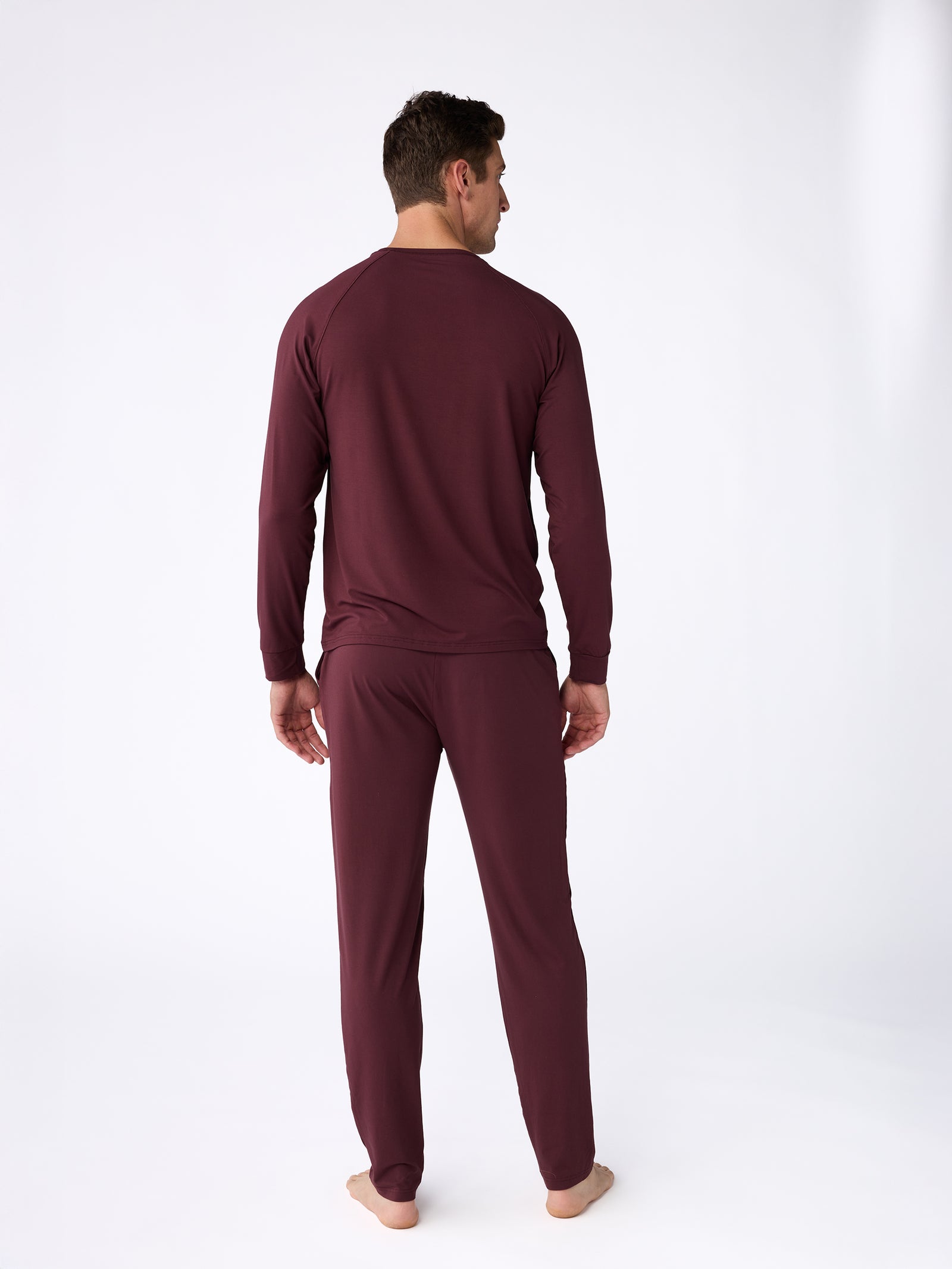 A man stands barefoot facing away, wearing the Men's Stretch-Knit Bamboo Long Sleeve by Cozy Earth and matching pants in dark maroon. The plain white background emphasizes the outfit's casual, comfortable style.