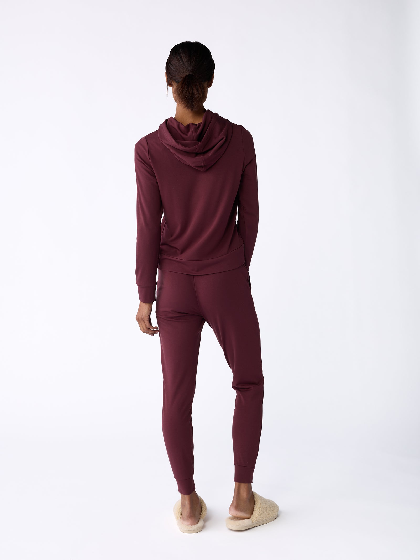 A person stands facing away, wearing Cozy Earth's Women's Bamboo Jogger Pant in maroon and beige slippers. The plain white background accentuates the outfit.