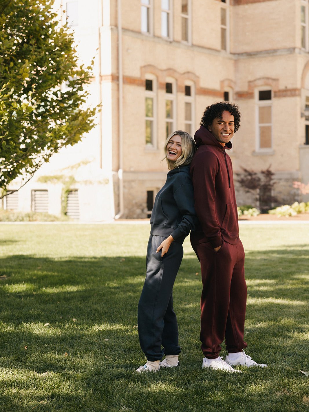 Two people stand back-to-back on a grassy lawn, both smiling. One is wearing a navy blue hoodie and a pair of Cozy Earth's Men's CityScape Sweatpants, while the other is dressed in a maroon hoodie and matching Men's CityScape Sweatpants from Cozy Earth. A tall, light-colored building and a tree can be seen in the background.