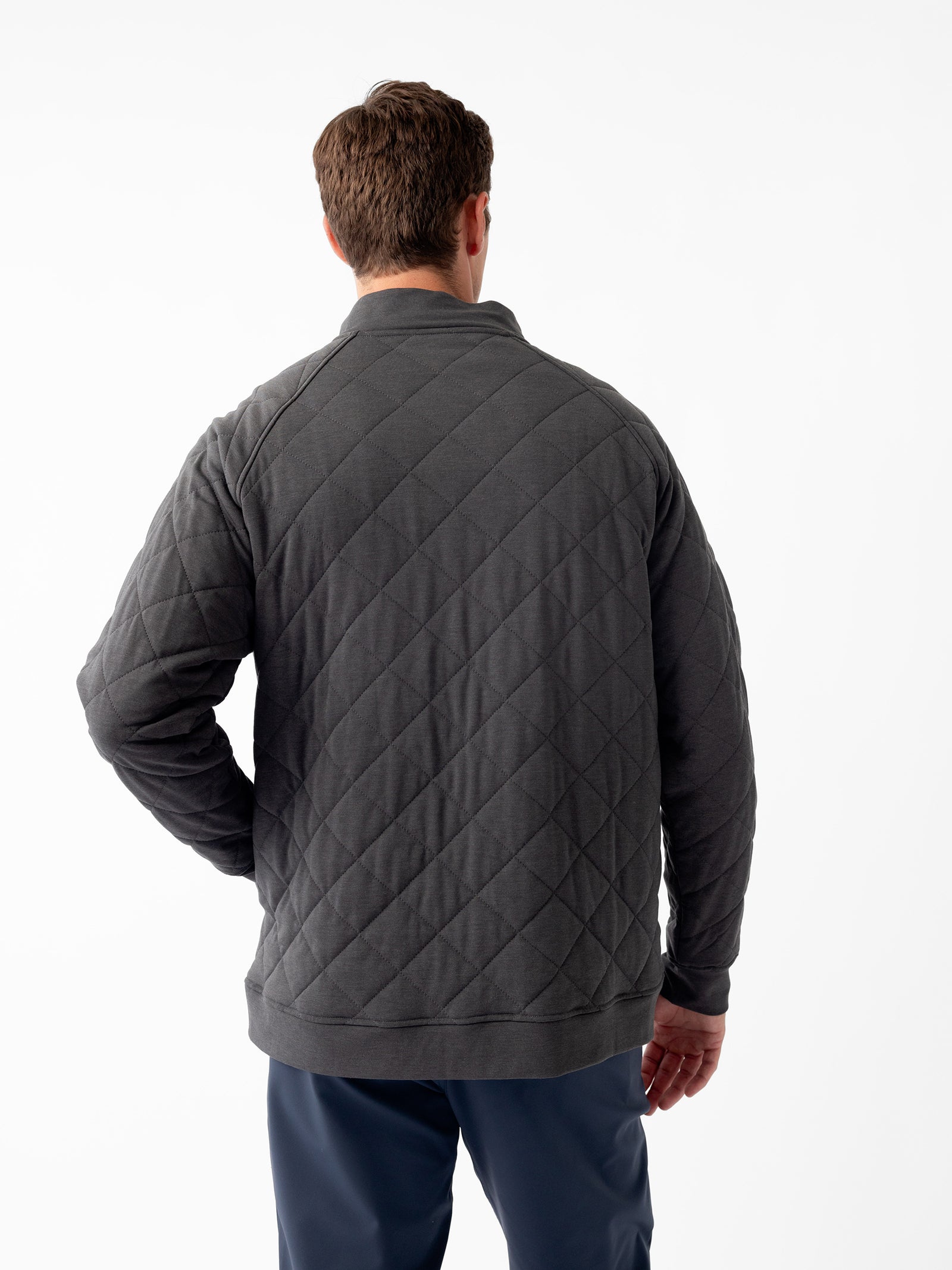 A man with brown hair is standing with his back to the camera, wearing a gray Men's Ultra-Soft Bamboo Quilted Snap Pullover from Cozy Earth and dark blue pants. His left hand is positioned on his hip. The background is plain white.
