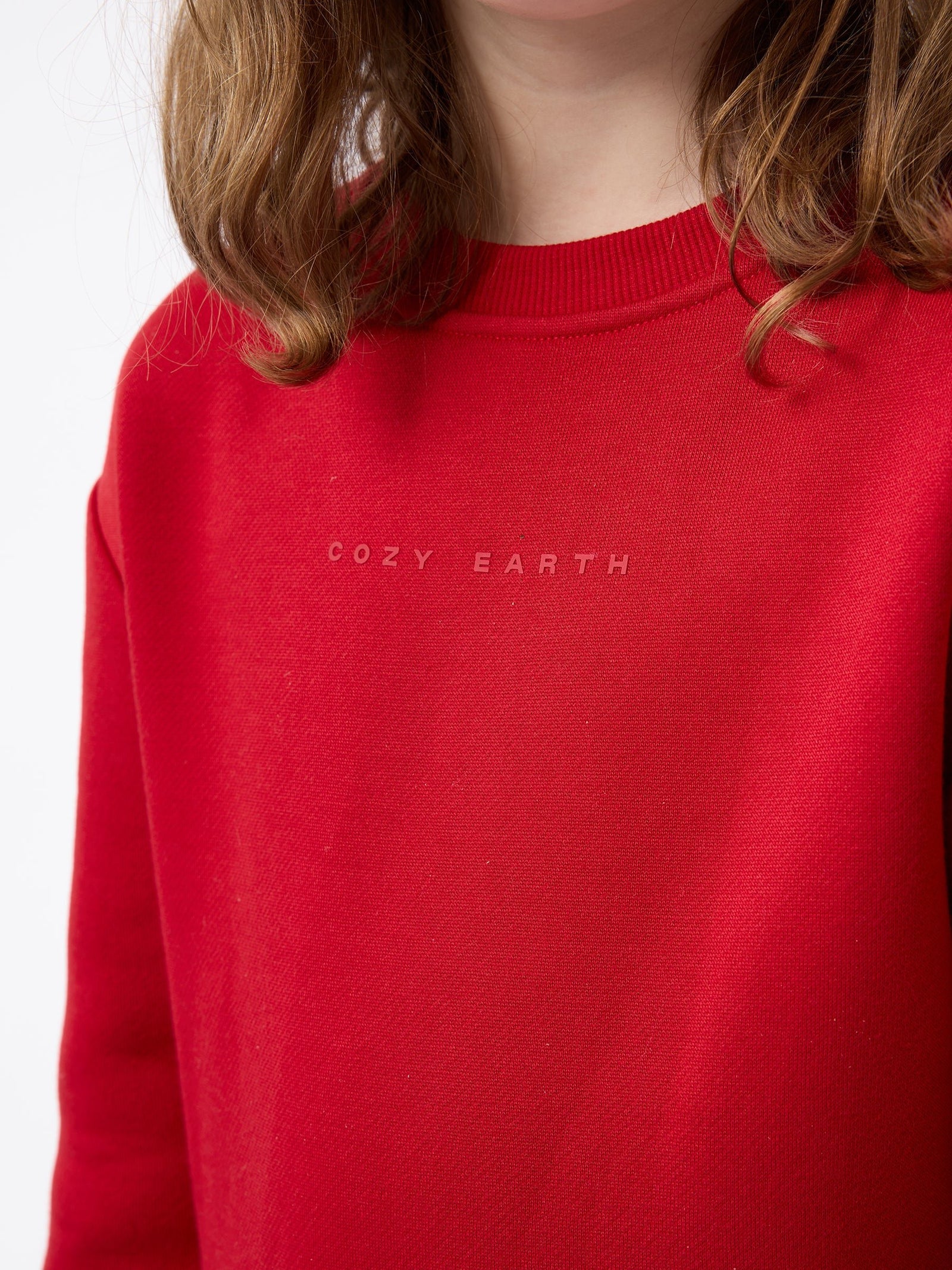 A person with shoulder-length hair wearing a red Kid's CityScape Crewneck by Cozy Earth. The background is plain white.