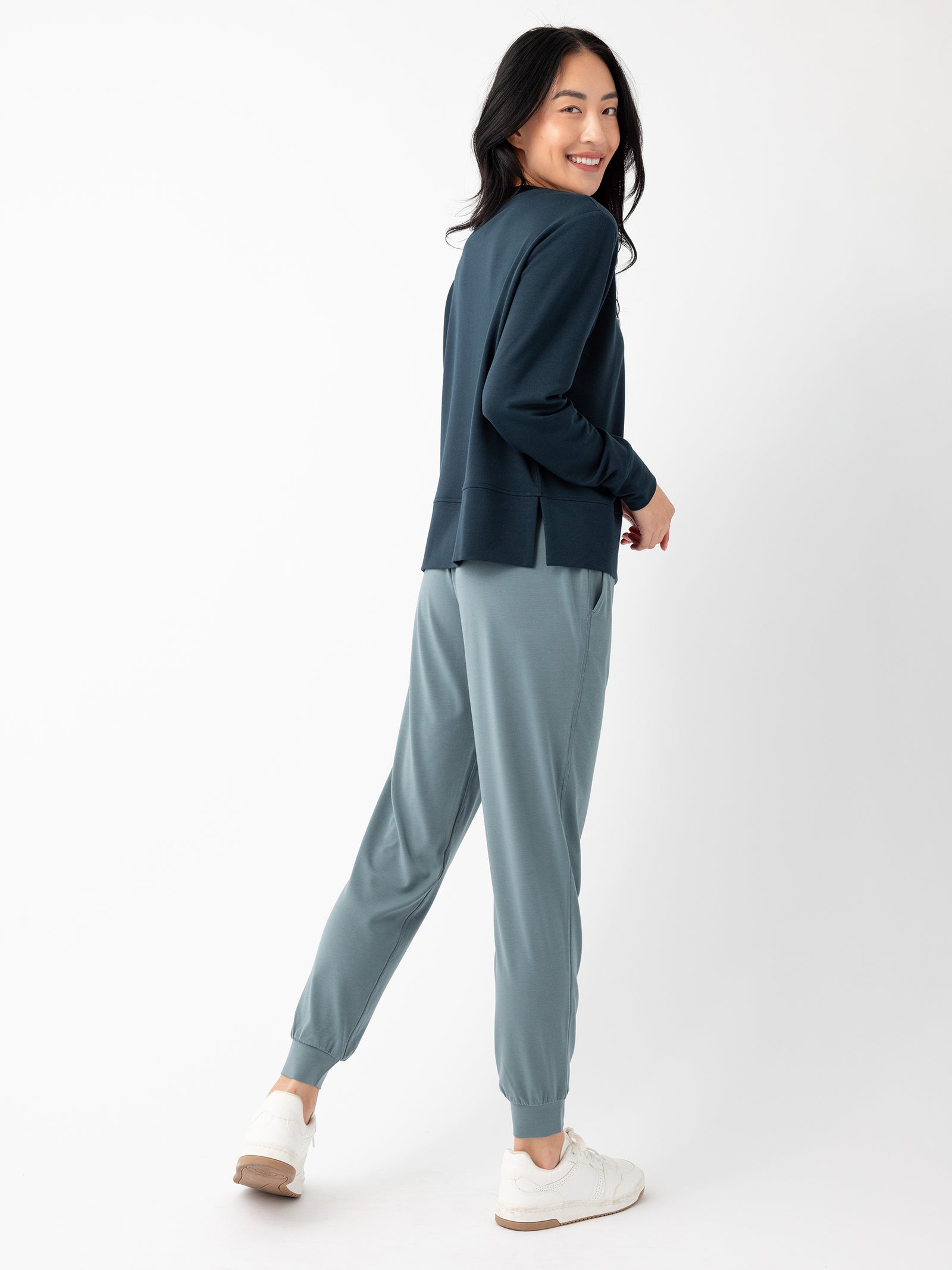 A person with long, dark hair smiles while posing in a dark green jacket and Cozy Earth's Women's Studio Jogger in light blue. They are standing against a plain white background and wearing white sneakers. Their body is turned slightly to the side, and they are looking back at the camera.