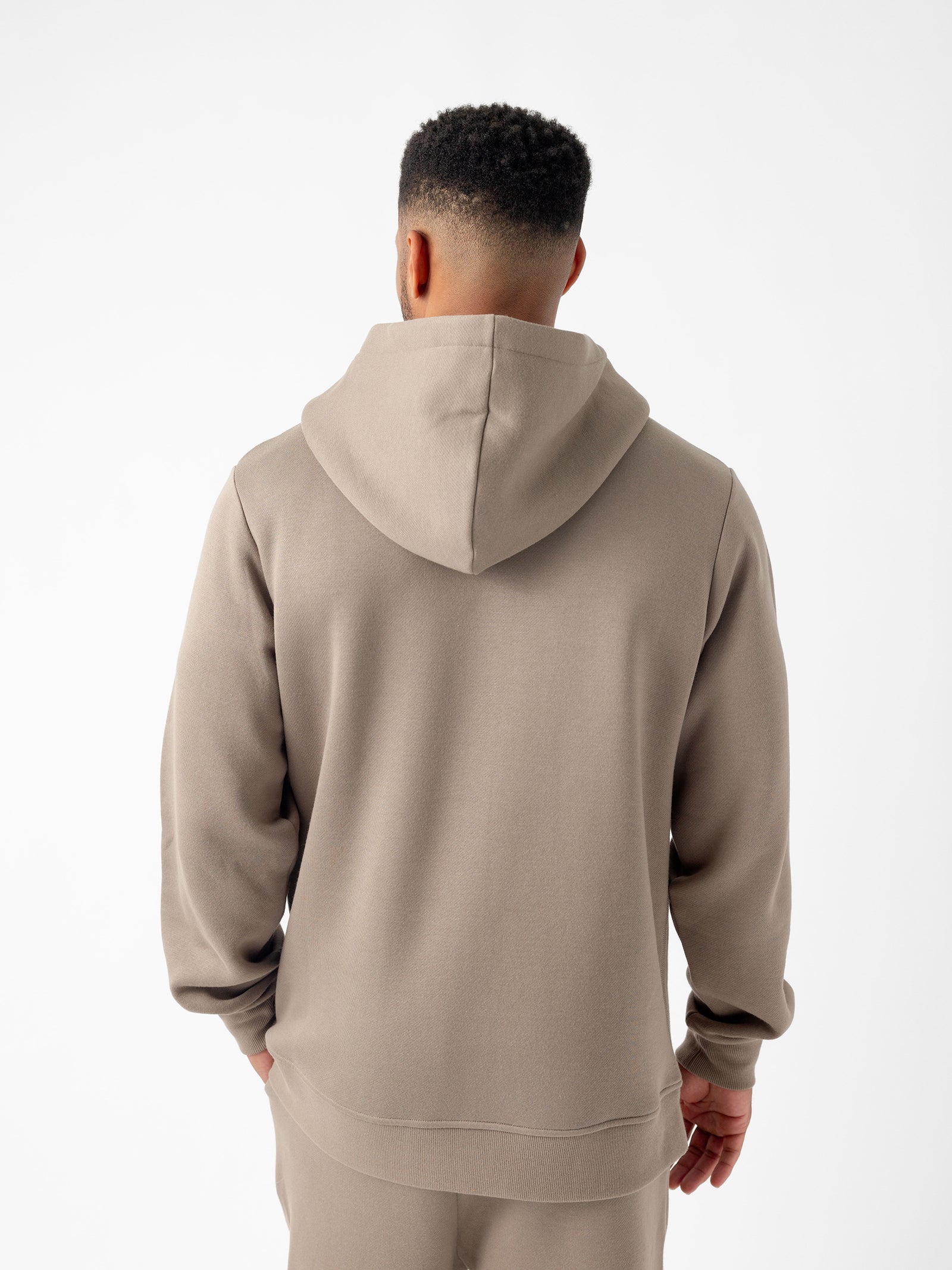 A person stands facing away from the camera, wearing a light brown Men's CityScape Hoodie and matching pants from Cozy Earth. The background is plain white, highlighting the outfit. The hoodie is plain with no visible designs or logos.