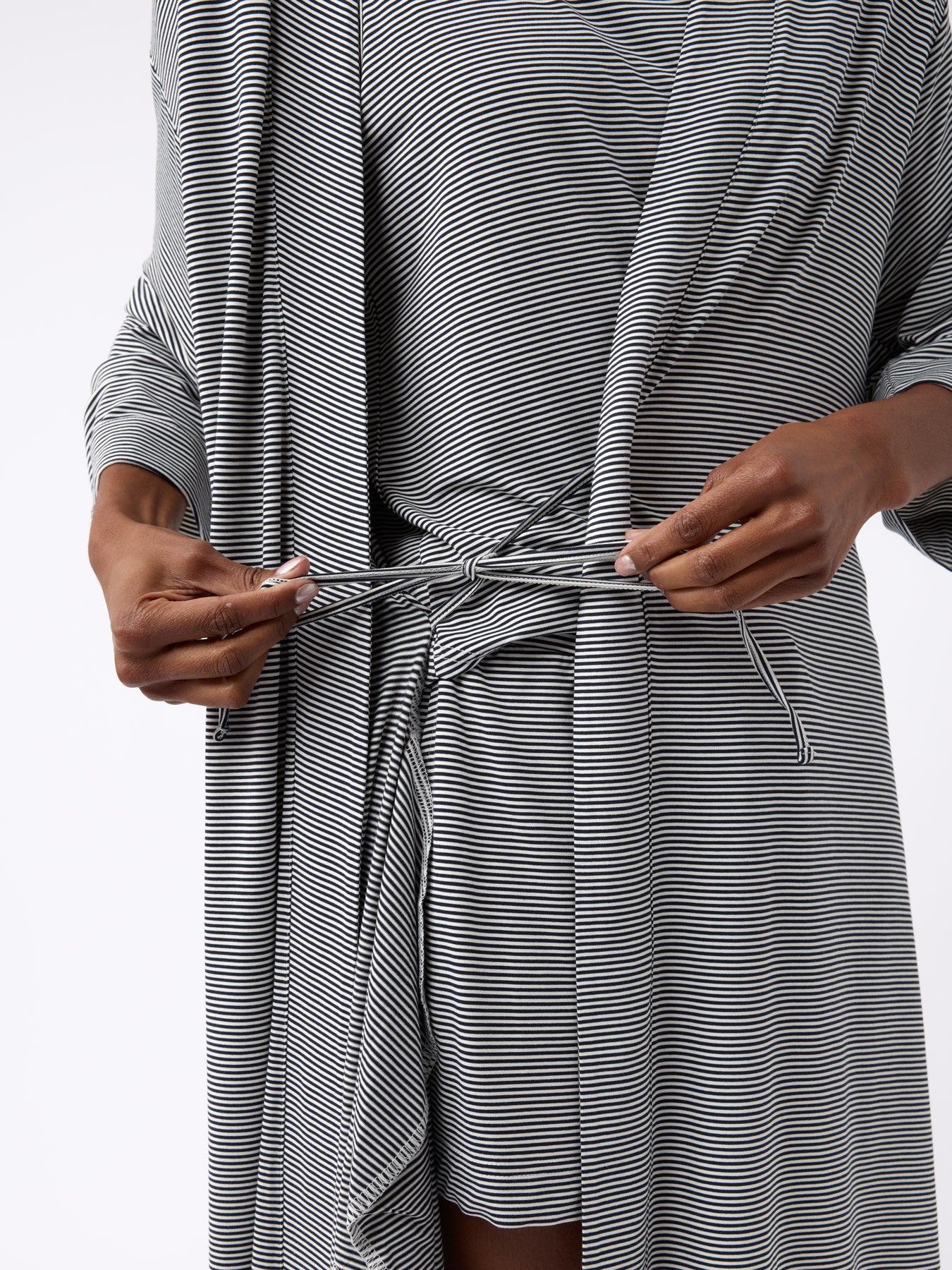 A person fastens the belt of a Cozy Earth Women's Bamboo Stretch-Knit Kimono Robe in black and white stripes. The lightweight robe has long sleeves, and the setting features a plain, light-colored background.