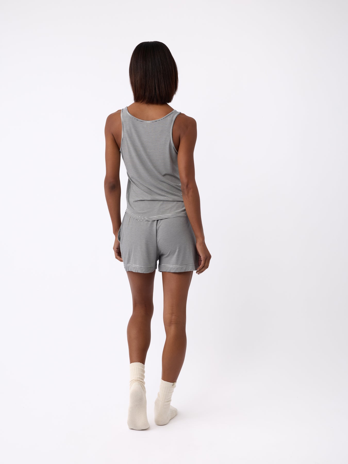 A person faces away, clad in a gray tank top, Cozy Earth's Women's Bamboo Stretch-Knit Shorts, and white socks. They sport short, straight hair against a plain backdrop.