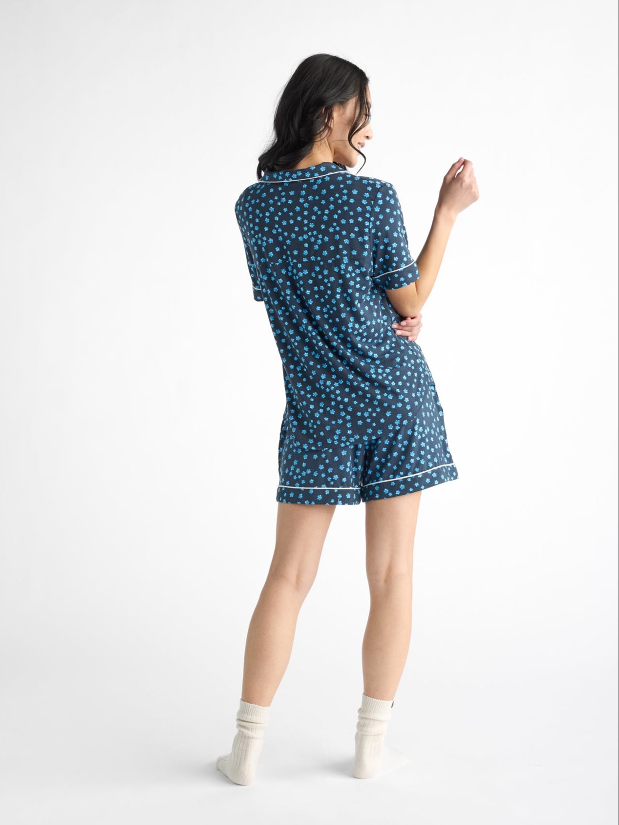 A person with shoulder-length dark hair stands barefoot, wearing white ankle socks and a blue Women's Bamboo Stretch-Knit Pajama Short from Cozy Earth, featuring a pattern of small, light blue dots. The background is plain white.