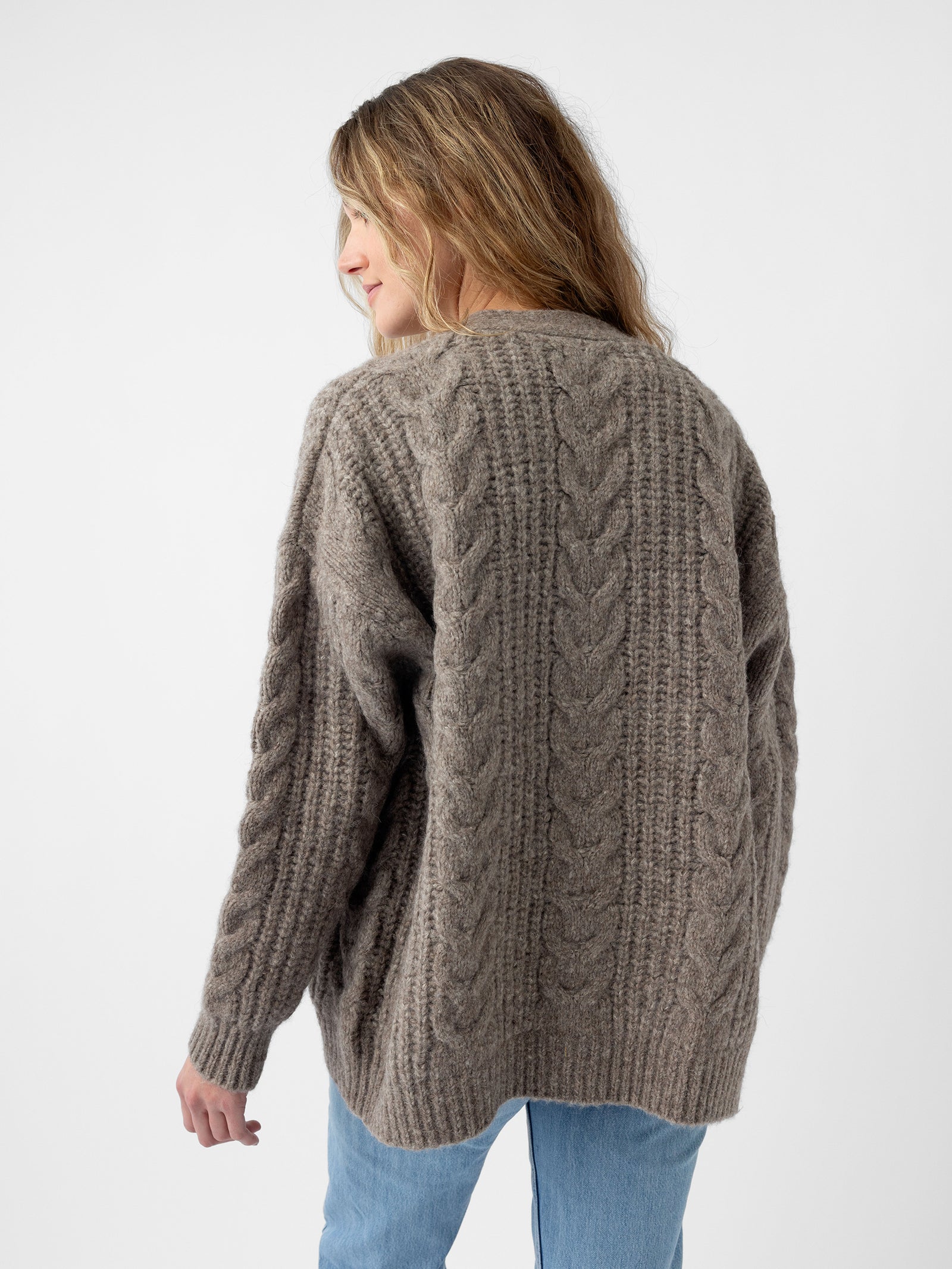 A person with long hair is wearing a grey Oversized Cable Knit Cardigan from Cozy Earth and blue jeans, standing against a plain white background with their head slightly turned to the side.