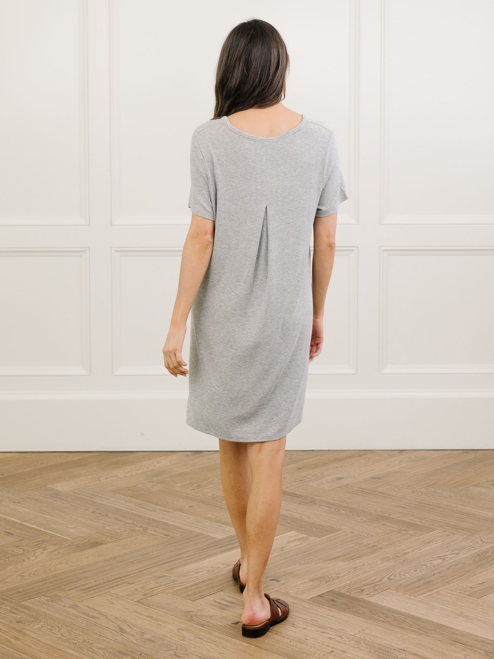 A woman facing backwards standing in a grey Cozy Earth bamboo rib-knit boyfriend sleep dress with short sleeves, knee-length, in a lounge or bedroom setting.