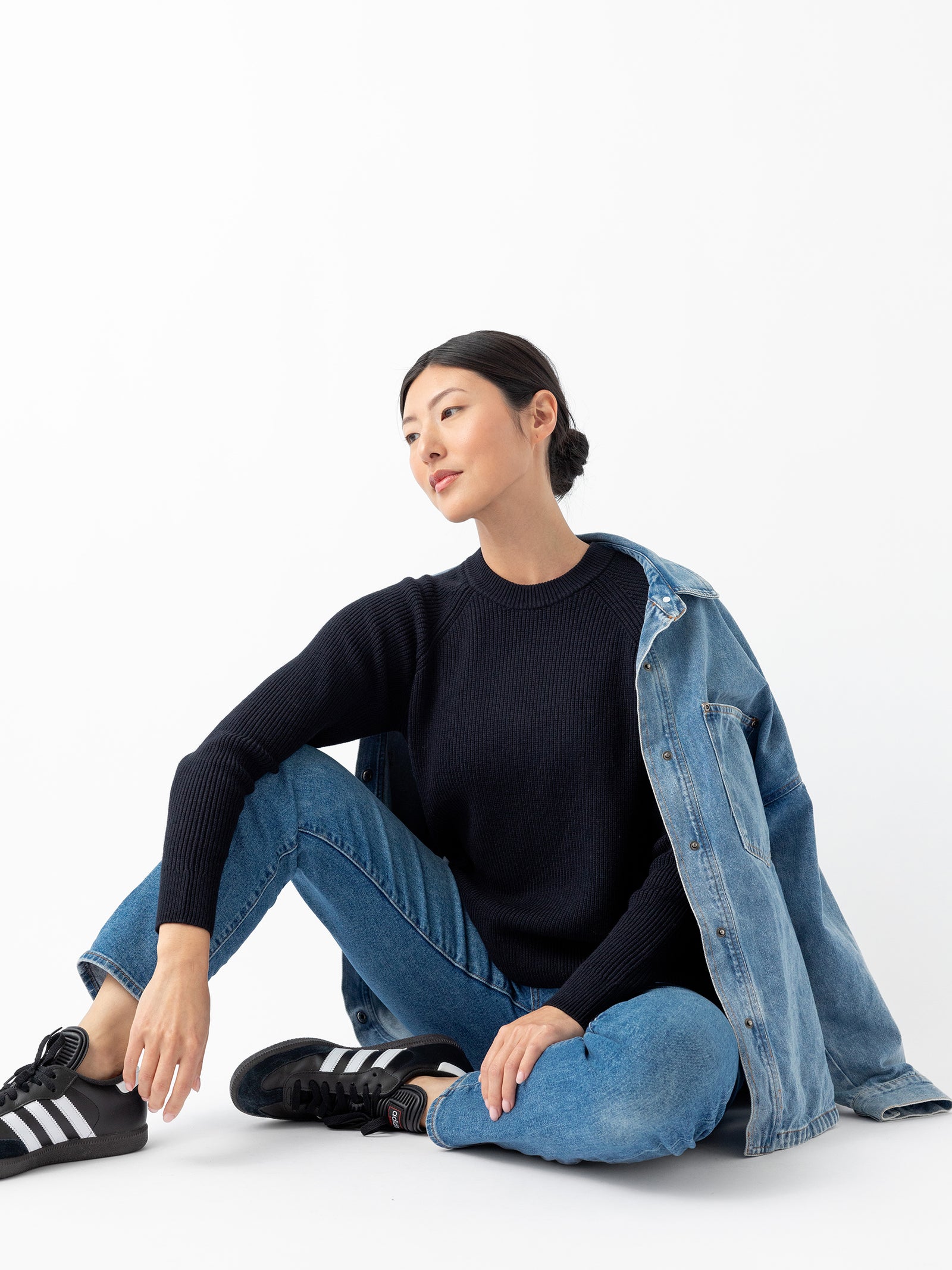 A person with long black hair, wearing Cozy Earth's Women's Classic Crewneck in black along with blue jeans and black sneakers featuring white stripes, sits on the floor with one leg bent. They have a denim jacket draped over their shoulder and gaze off to the side against a plain white background.