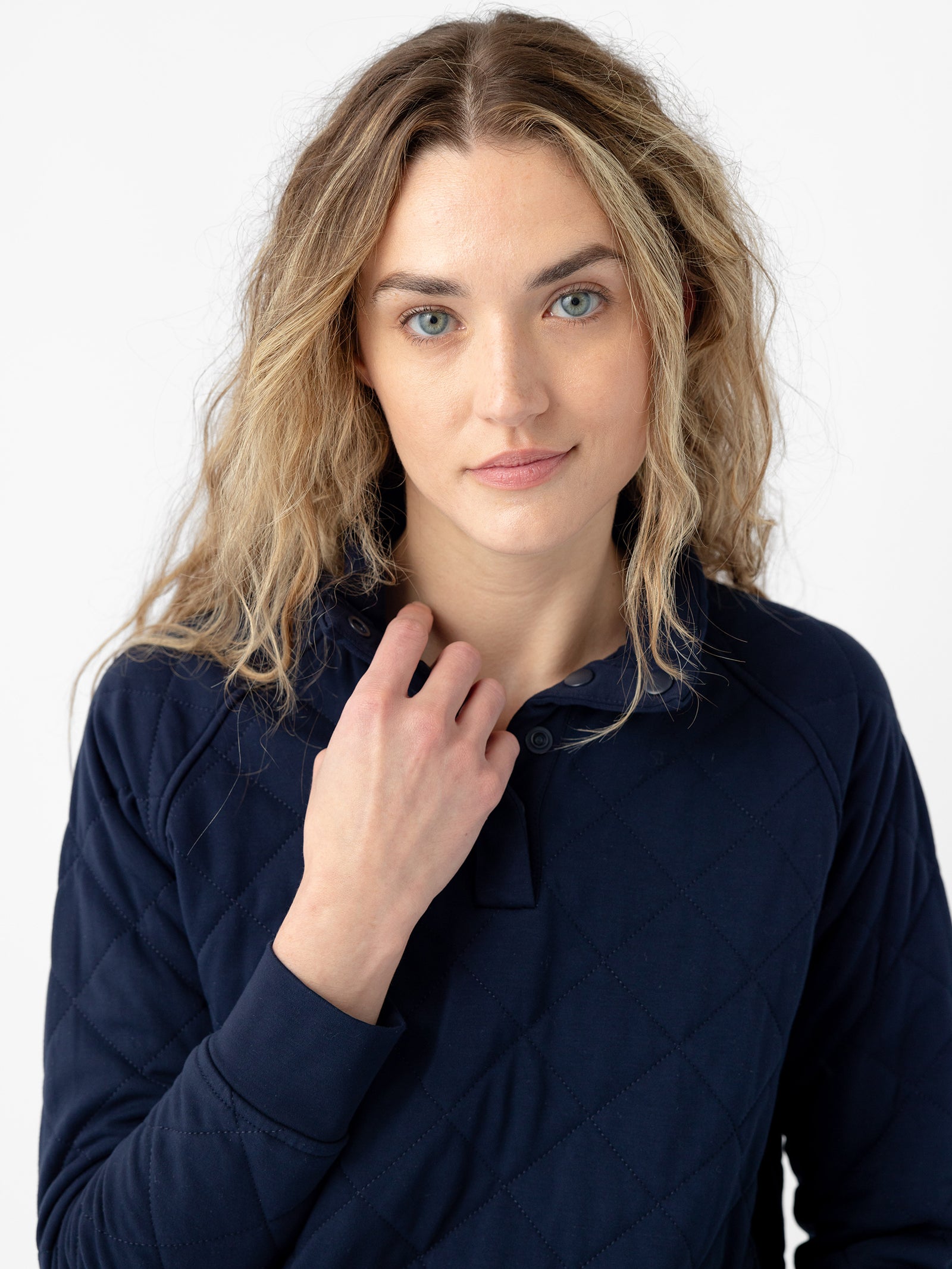 A person with long, wavy blonde hair is wearing a Women's Ultra-Soft Bamboo Quilted Snap Pullover from Cozy Earth. They are gazing calmly at the camera, with one hand raised near their collar. The background is plain white.