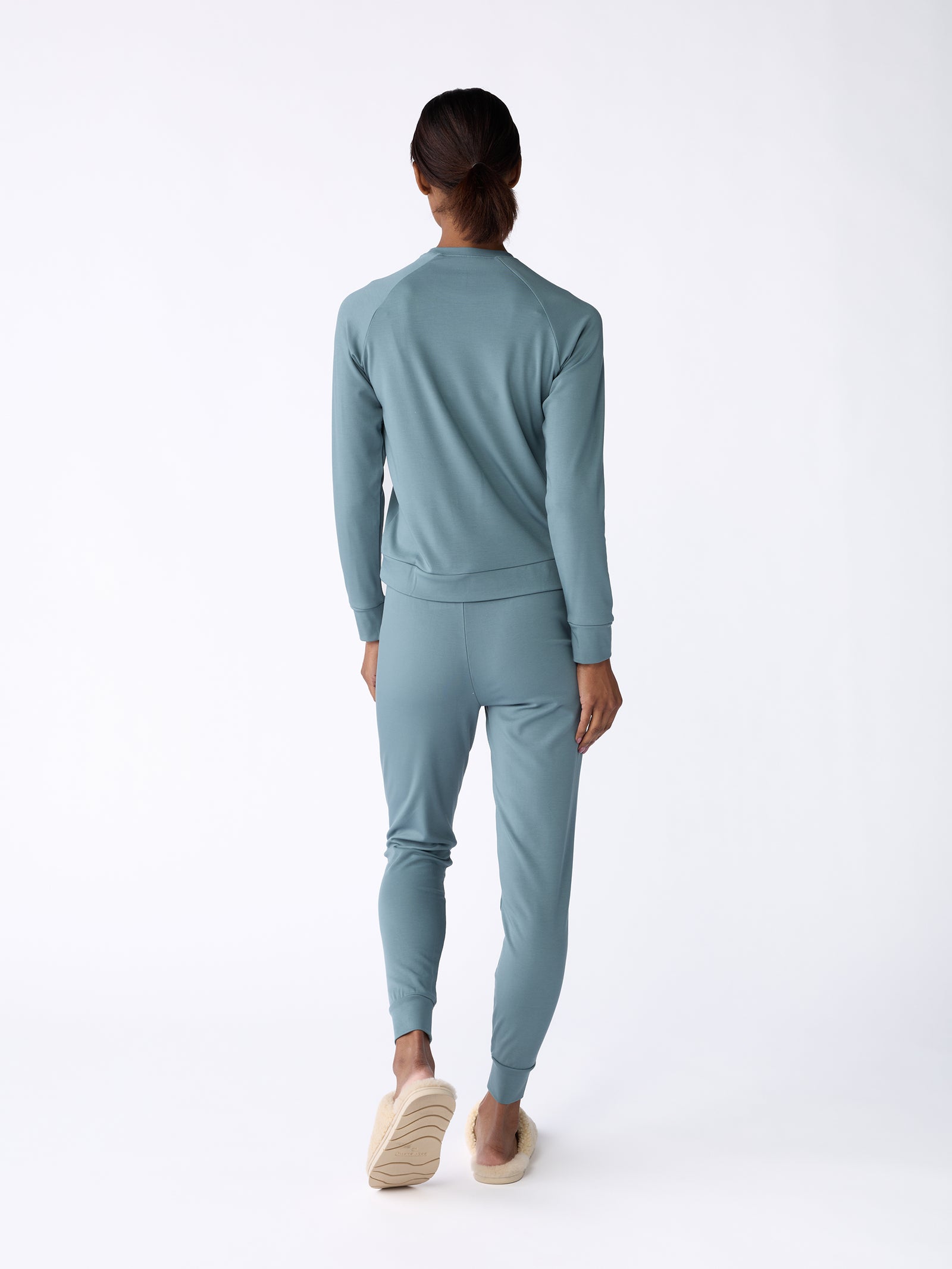 A woman is seen facing away, dressed in a matching blue-green tracksuit, featuring the Women's Bamboo Jogger Pant by Cozy Earth, complemented by beige slip-on shoes. The plain white background highlights the clothing ensemble.