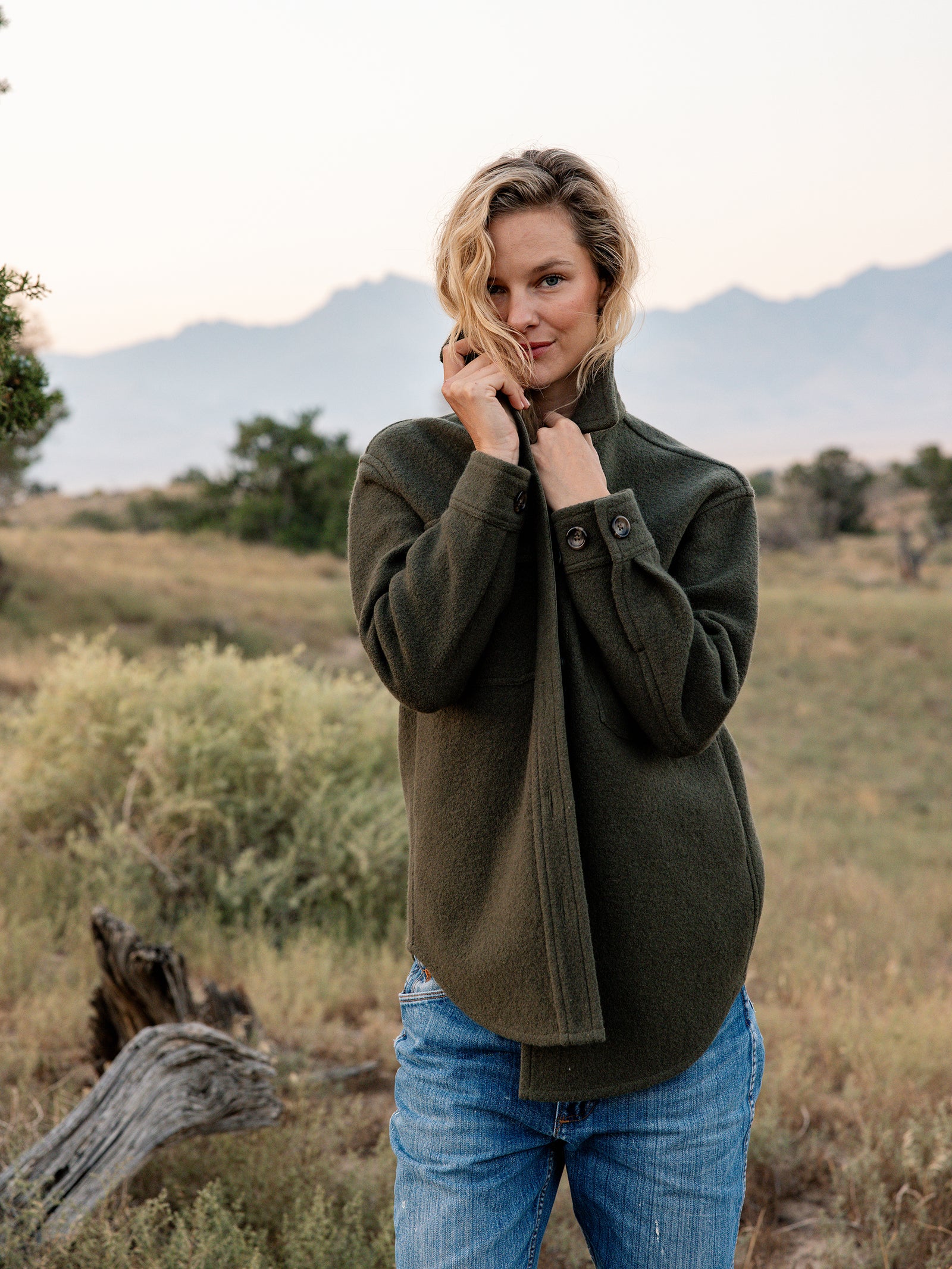A person with wavy blond hair stands outdoors in a natural setting, wearing the green Women's Boucle Shacket by Cozy Earth and blue jeans. They appear to be holding the collar of their shacket with one hand. The background features trees, bushes, and distant mountains.