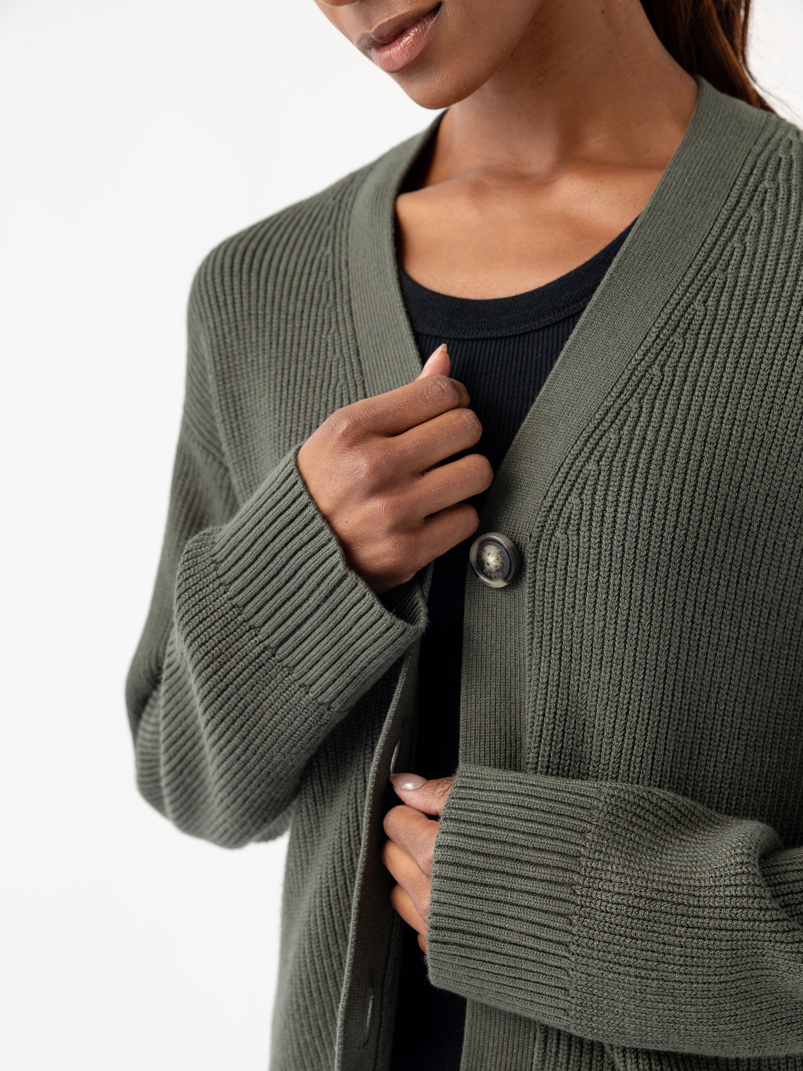 A woman is wearing a Cozy Earth Oversized Classic Cardigan in green over a black top. The cardigan features a prominent button that is fastened at the top, and the image highlights her upper body and hands against a plain white background.