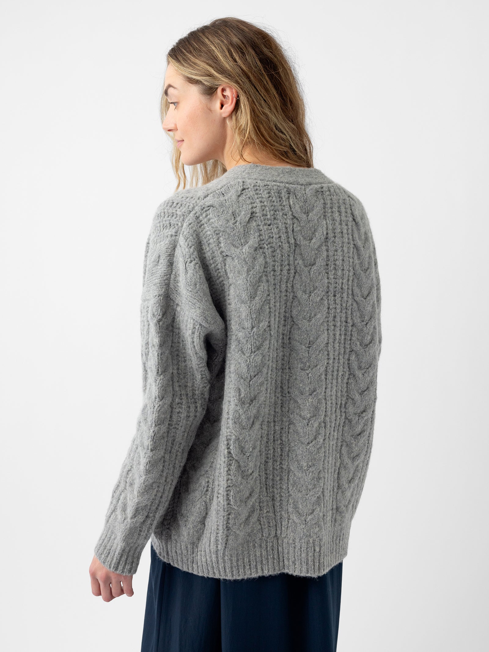 A woman with long hair is wearing a gray Oversized Cable Knit Cardigan from Cozy Earth. She is facing away, with her head turned slightly to the side, against a plain white background. The bottom of her skirt peeks into view at the edge of the image.