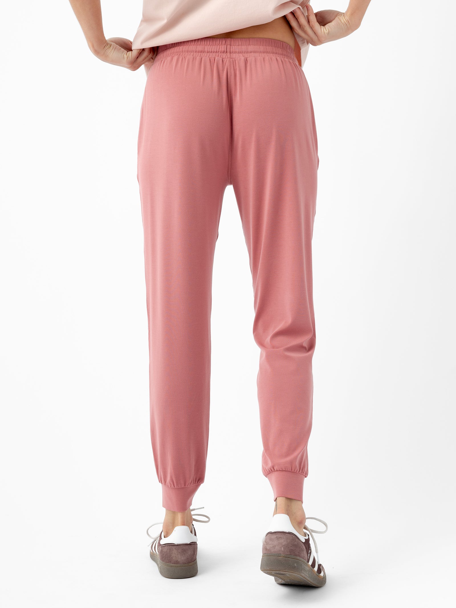 A person standing with their back to the camera, wearing Cozy Earth's rose-colored Women's Studio Jogger and an off-white top. They are also wearing brown and white sneakers. The person's hands are positioned on their hips against a plain white background.