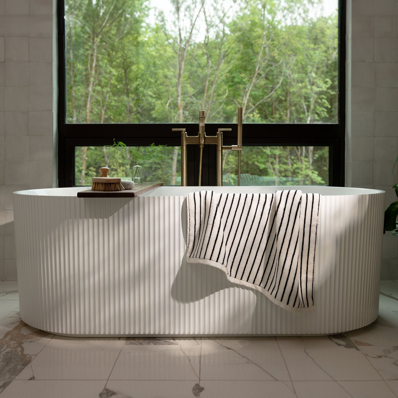 A serene bathroom features a white freestanding bathtub with a Luxe Bath Towel from Cozy Earth draped over the side. Behind the tub, a large window showcases a lush green forest, allowing natural light to flood the space. The floor is made of white marble with subtle grey veins.