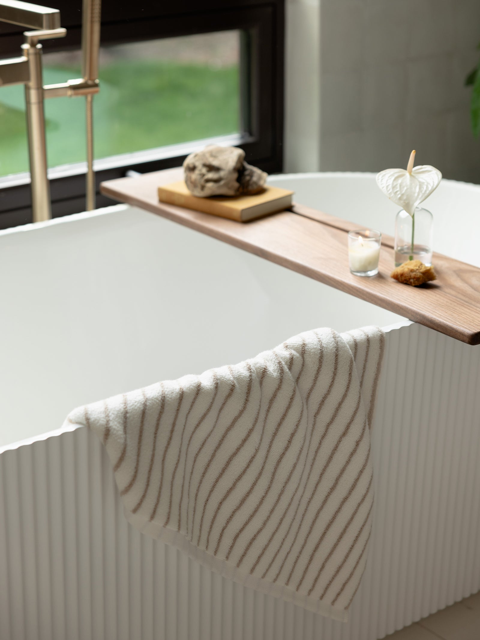 A modern white bathtub with a textured exterior stands near a window. A wooden tray spans the tub, holding two glass vases with plants, a candle, and a decorative stone. Draped over the tub's edge is a Luxe Bath Towel featuring thin, wavy brown stripes from Cozy Earth.