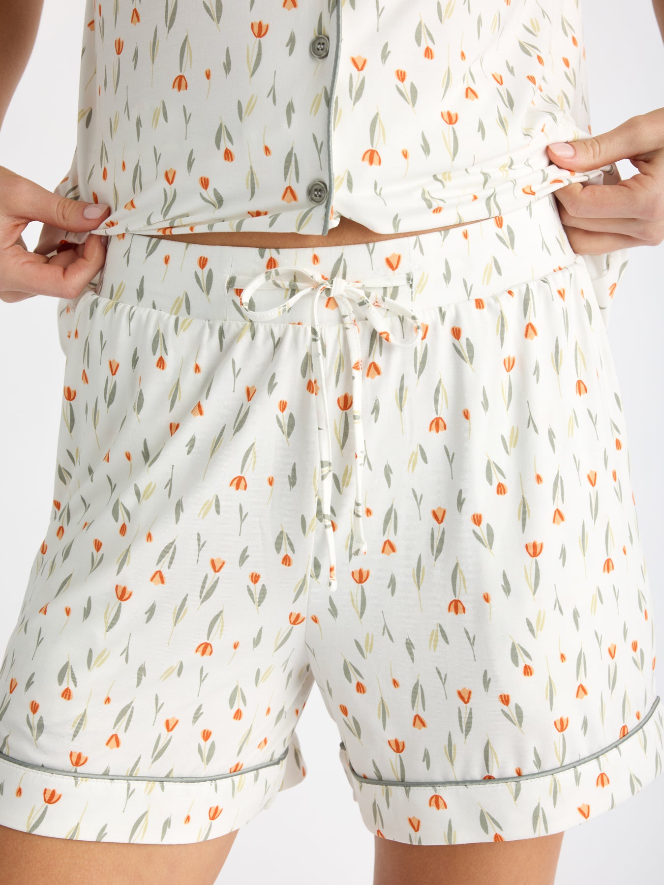 A person wearing HIDE's Women's Bamboo Stretch-Knit Pajama Short in a white floral pattern with small orange tulips adjusts their button-up top, revealing the waistband of the shorts.