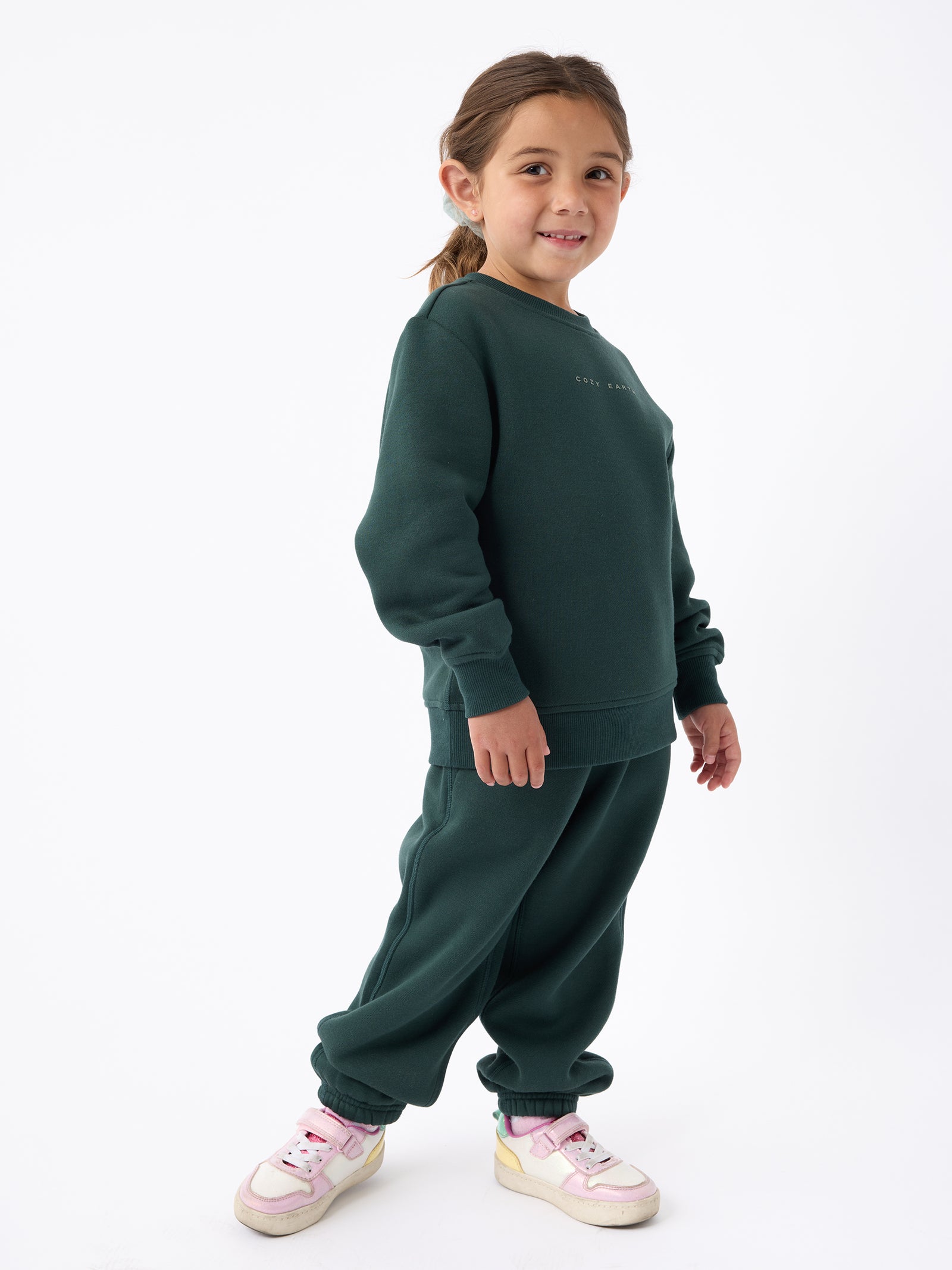 A young child with brown hair stands against a white background, smiling. They are wearing a dark green Kid's CityScape Crewneck by Cozy Earth, paired with sweatpants and pink and white sneakers.