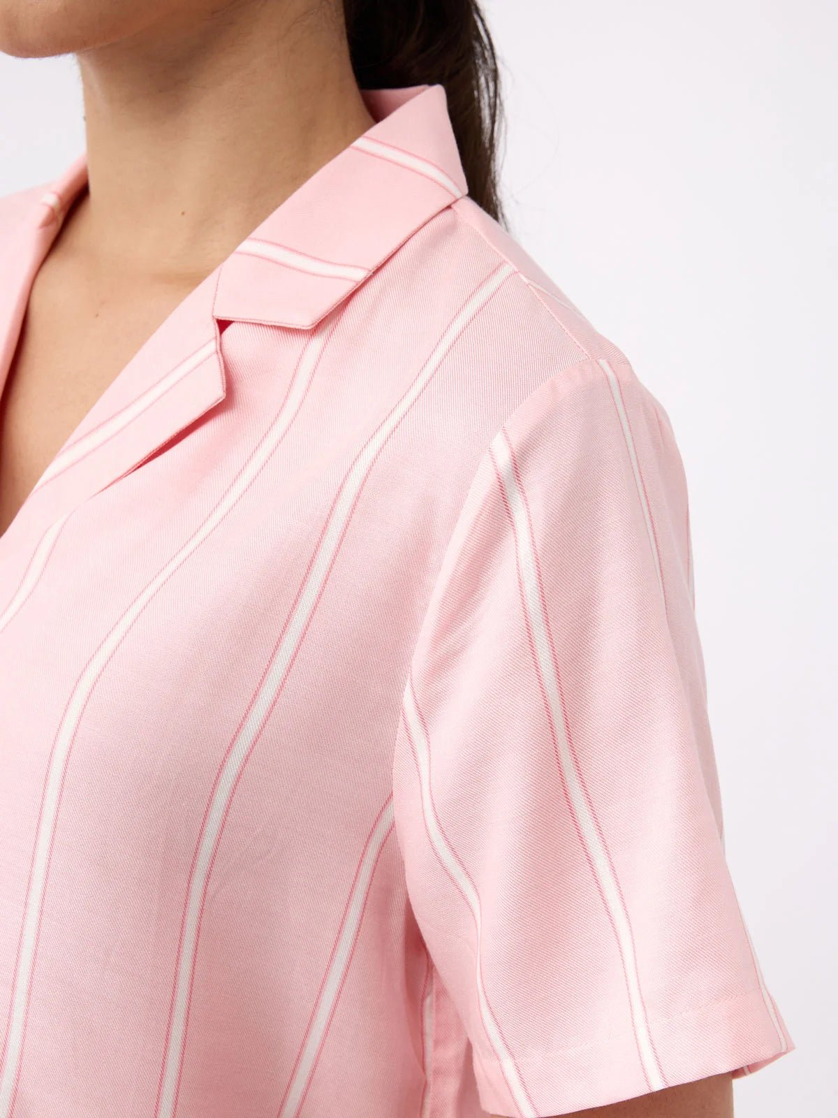 A close-up shows a person wearing the Women's Soft Woven Short Sleeve Pajama Top by Cozy Earth, featuring a pink color with white stripes, collar, and short sleeves against a plain white background.