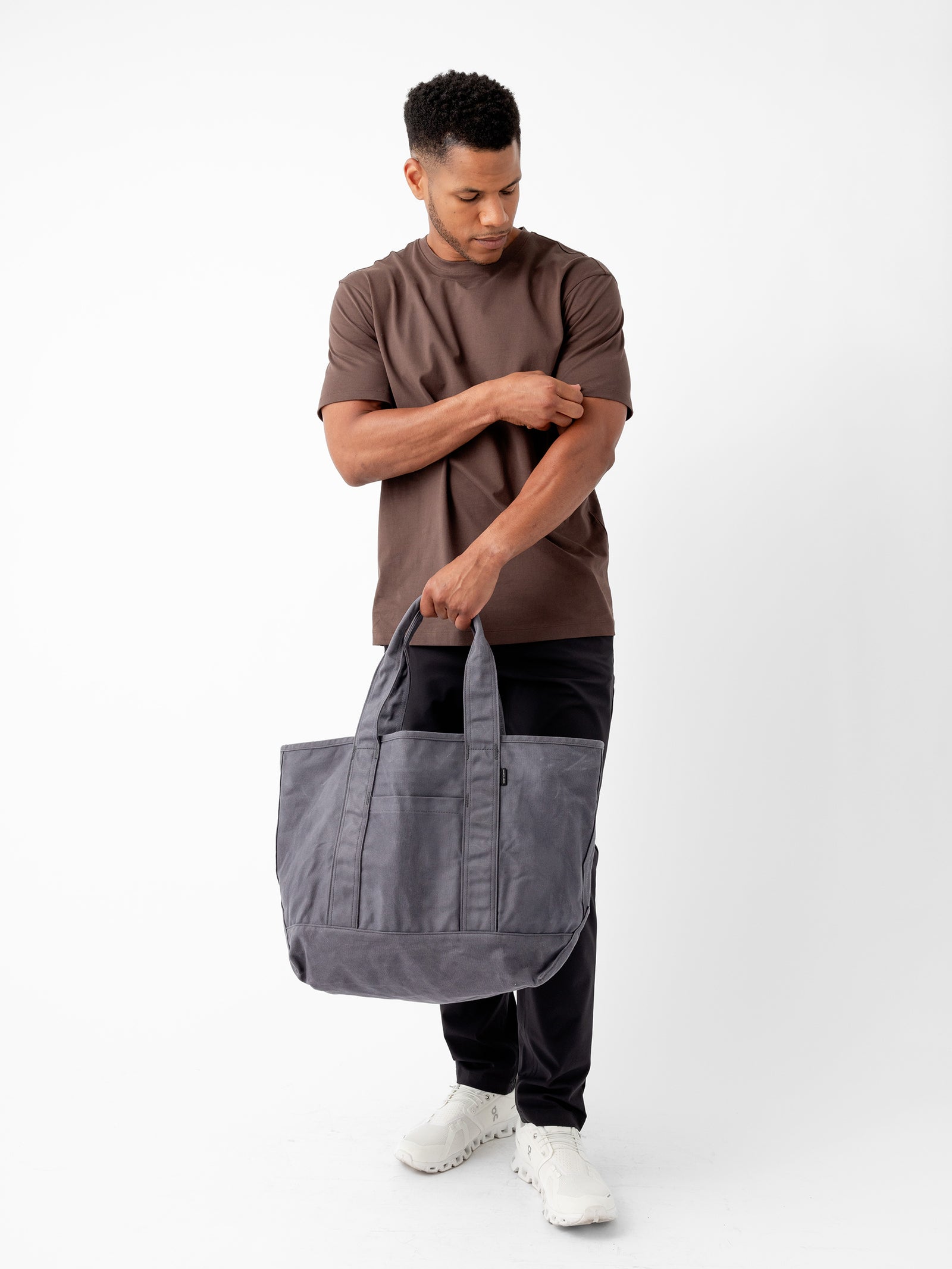 A man with short hair stands against a plain white background, looking down as he adjusts the sleeve of his brown T-shirt. In his left hand, he holds a large Waxed Canvas Tote by Cozy Earth, featuring multiple pockets. He is also wearing black pants and white sneakers.