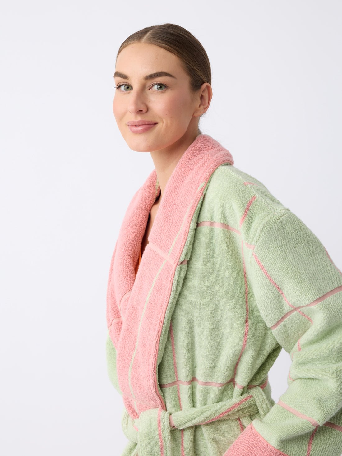 A woman with slicked-back hair is smiling and standing against a plain white background while wearing the Windowpane Resort Robe by Cozy Earth, featuring a pastel green color with pink accents, a checkered pattern, and pink collar and belt.