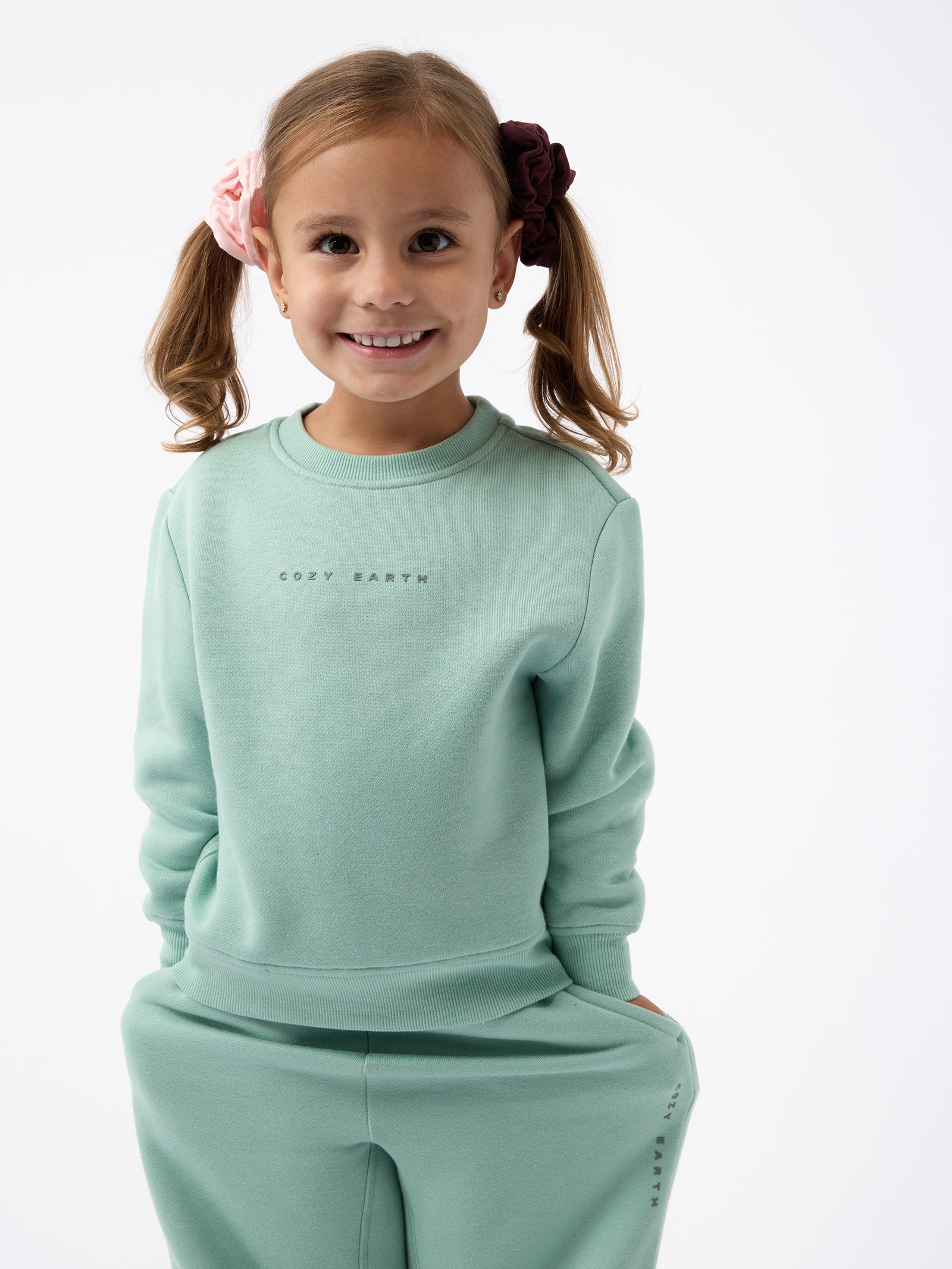 A young girl with ponytails, each tied with a colorful scrunchie, wears the mint green Kid's CityScape Crewneck and matching pants. She smiles at the camera against a plain white background, with "Cozy Earth" showcased on her sweatshirt.
