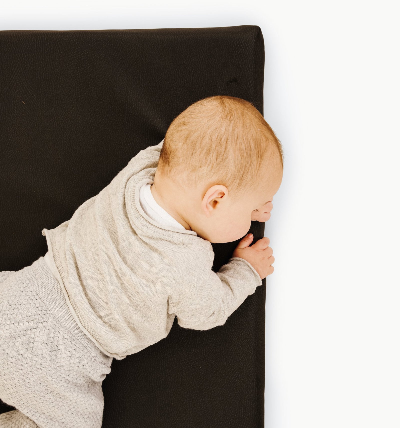A baby in a beige outfit lies on their stomach on a Gathre Wholesale Padded Changing Mat, head turned to the side. The scene is set against a white, minimalistic background.