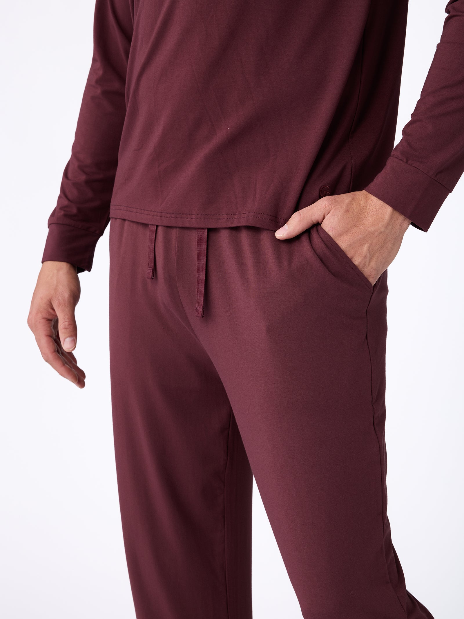 A person wearing a matching maroon long-sleeve shirt and the Men's Stretch-Knit Bamboo Pajama Pant by Cozy Earth. The outfit is casual with a drawstring waistband, and the person's right hand is in the pants pocket. The background is plain white.
