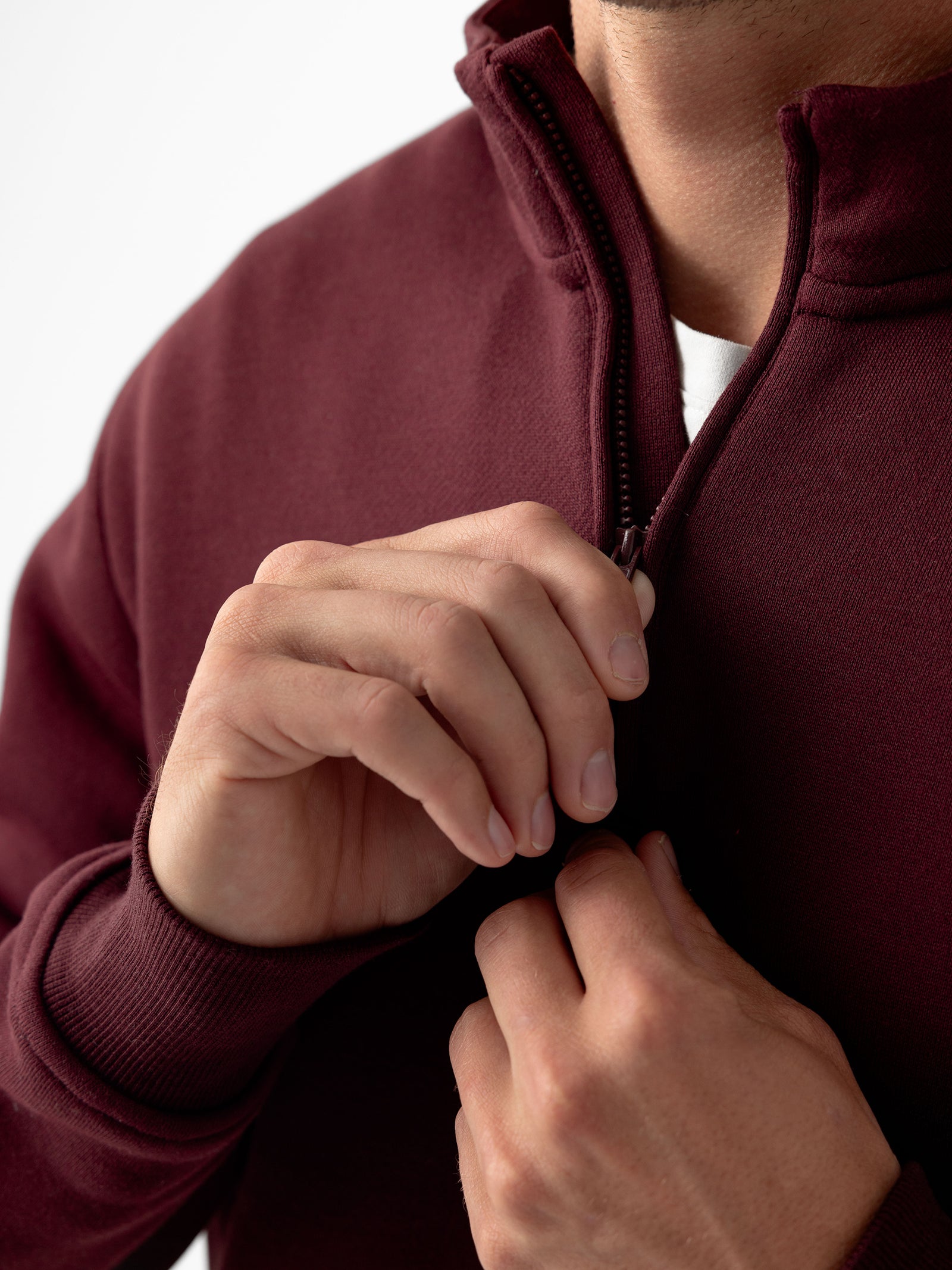 Close-up of a person zipping up a Cozy Earth Men's CityScape Quarter Zip sweater. The person's hands are visible, one holding the zipper pull while the other steadies the maroon fabric. The background is plain white.