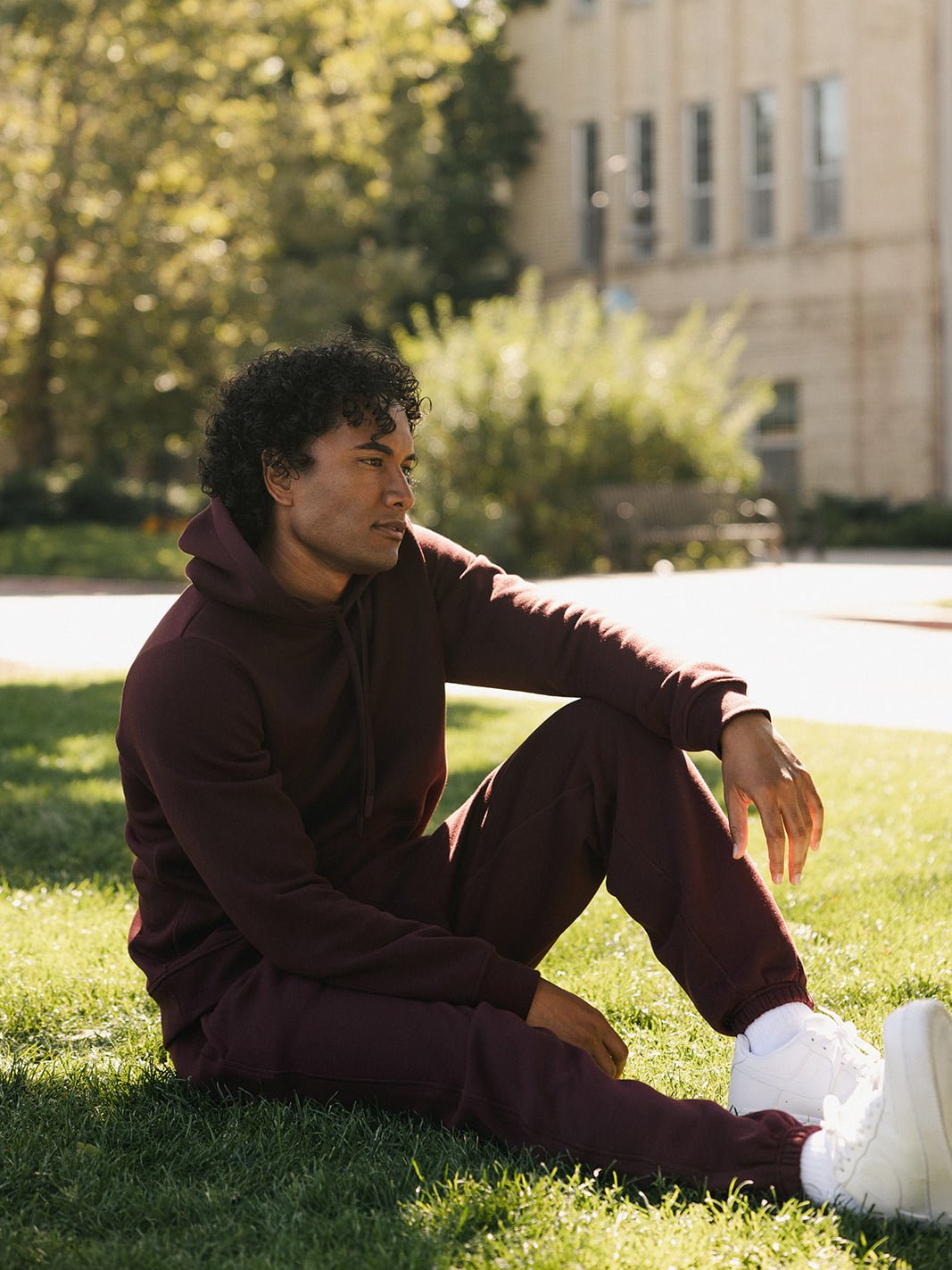 A person with curly hair is sitting on grassy ground in a relaxed pose, wearing a maroon hoodie, Men's CityScape Sweatpant by Cozy Earth, and white sneakers. The background features a tree-lined pathway and a building with large windows, bathed in sunlight.
