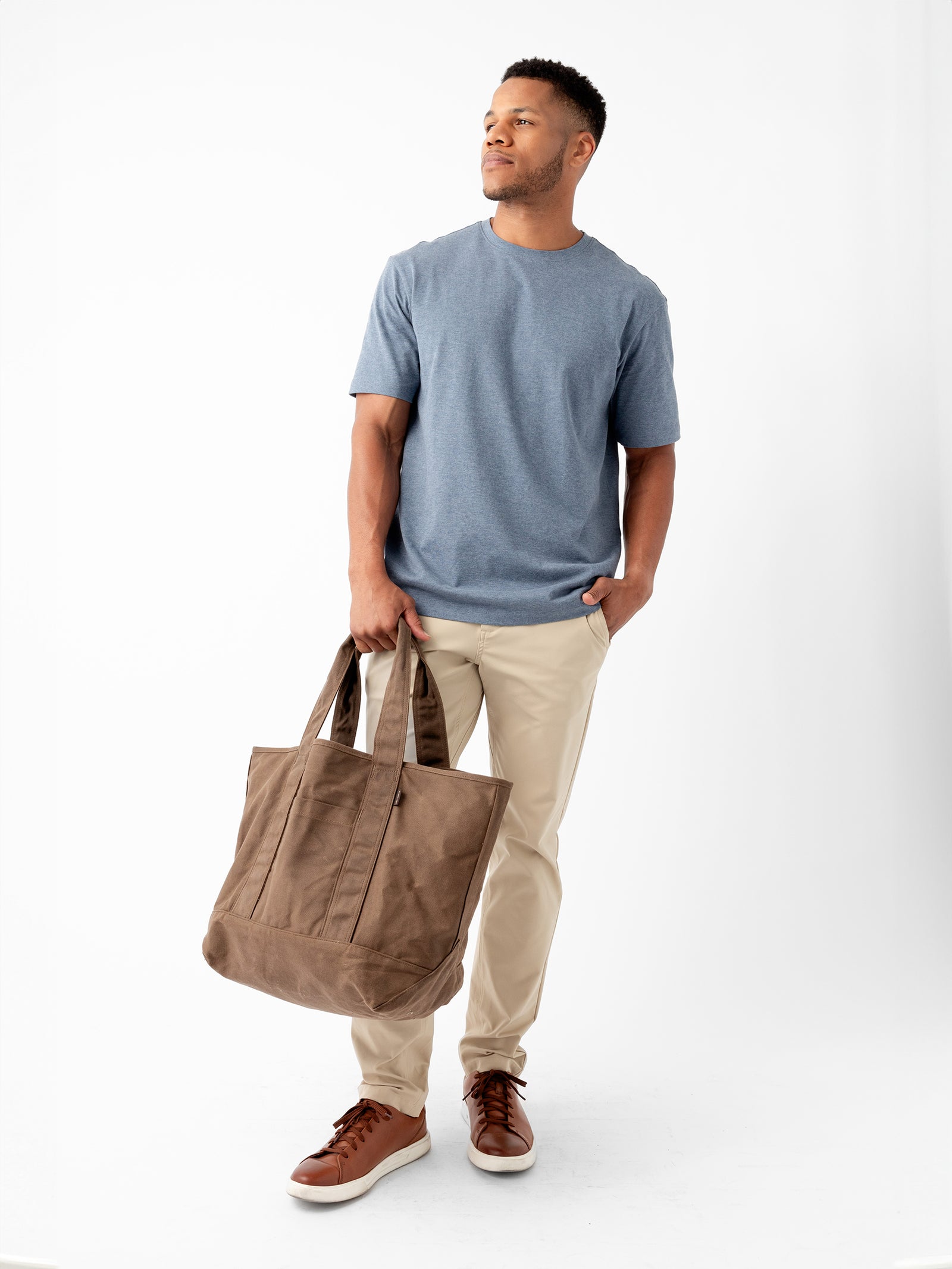 A man stands against a plain white background. He is wearing a blue T-shirt, beige pants, and brown sneakers. He holds a large Waxed Canvas Tote from Cozy Earth in his left hand and has his right hand in his pants pocket. He looks slightly to the left with a neutral expression.