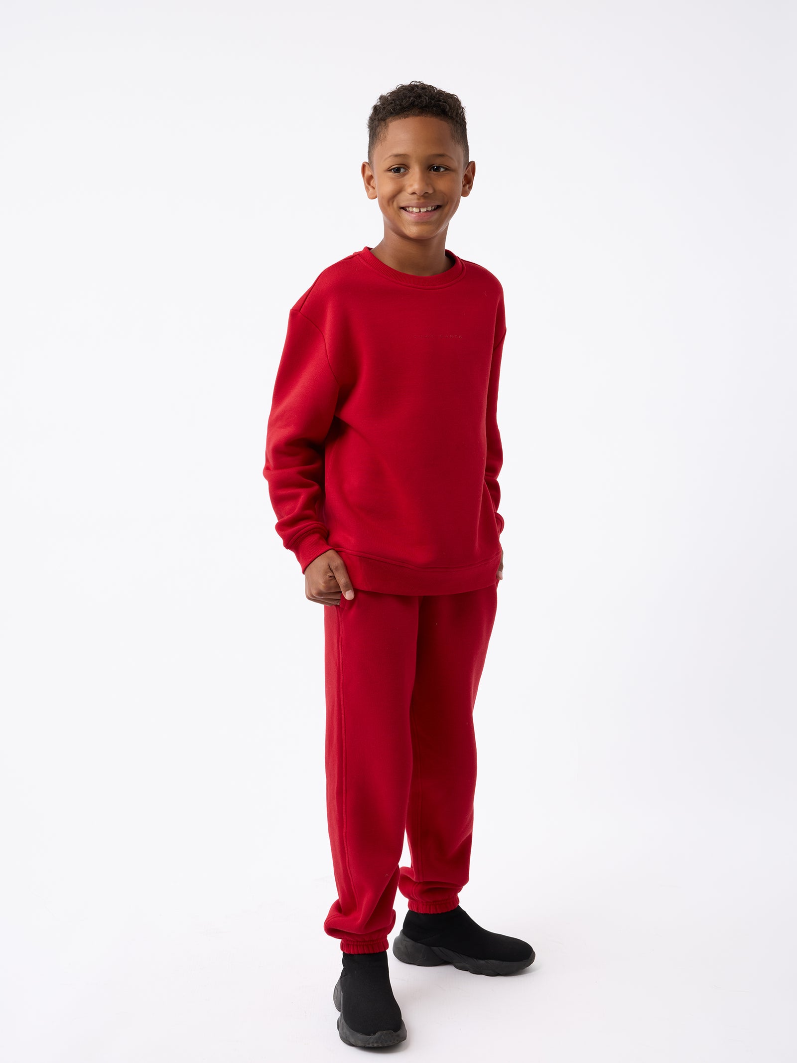 A boy wearing the Kid's CityScape Crewneck by Cozy Earth and black shoes stands smiling against a plain white background.