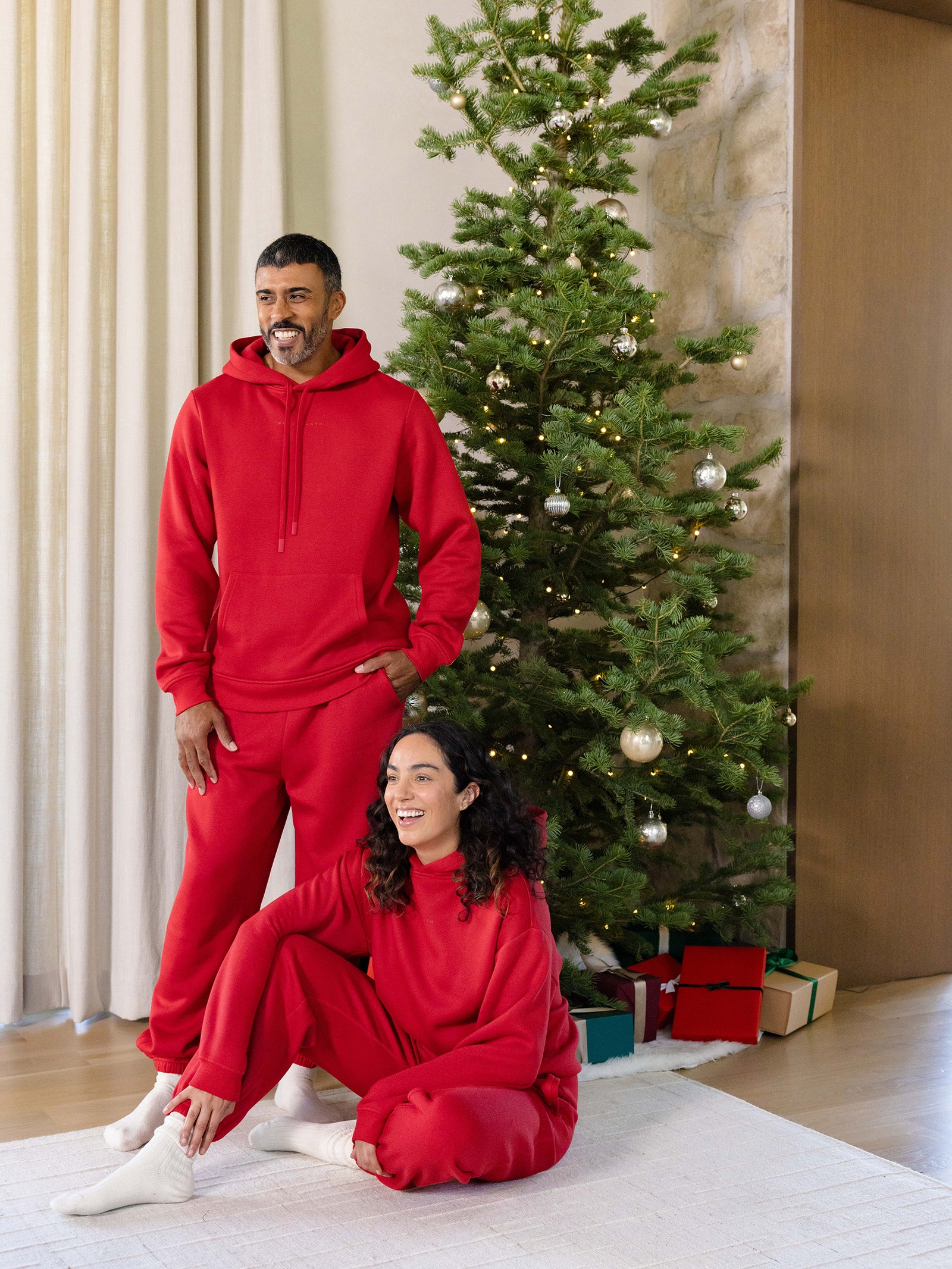 A man and a woman wearing matching Men's CityScape Hoodies from Cozy Earth are in a cozy living room decorated for Christmas. The woman sits on the floor, smiling, while the man stands behind her. A Christmas tree adorned with ornaments and presents is visible in the background.