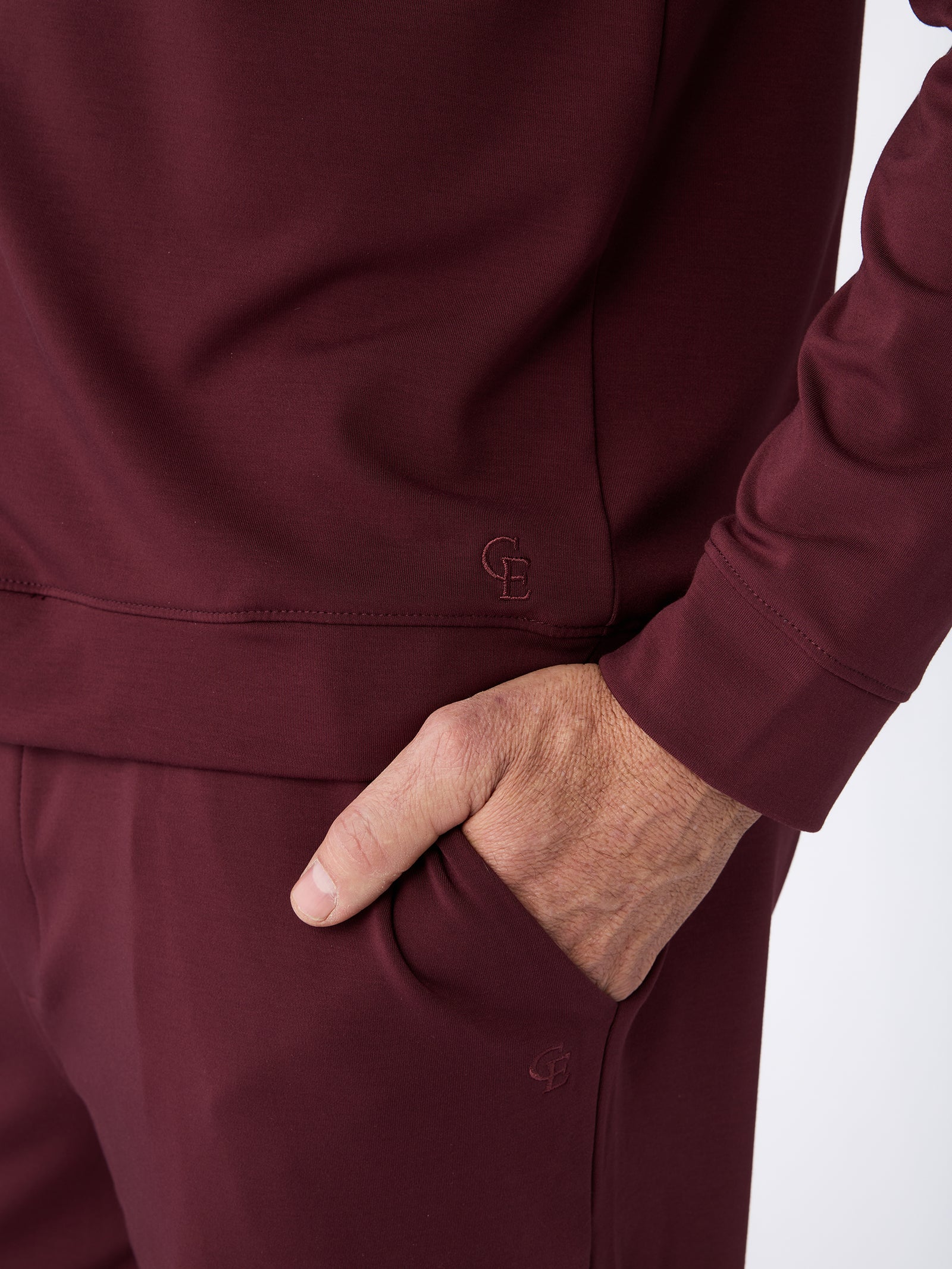 A close-up of a person wearing a burgundy Men's Ultra-Soft Bamboo Pullover Crew from Cozy Earth. The person's right hand is resting in the pocket of the pants, and the outfit includes subtle branding with "CB" initials embroidered near the waistband and pocket.