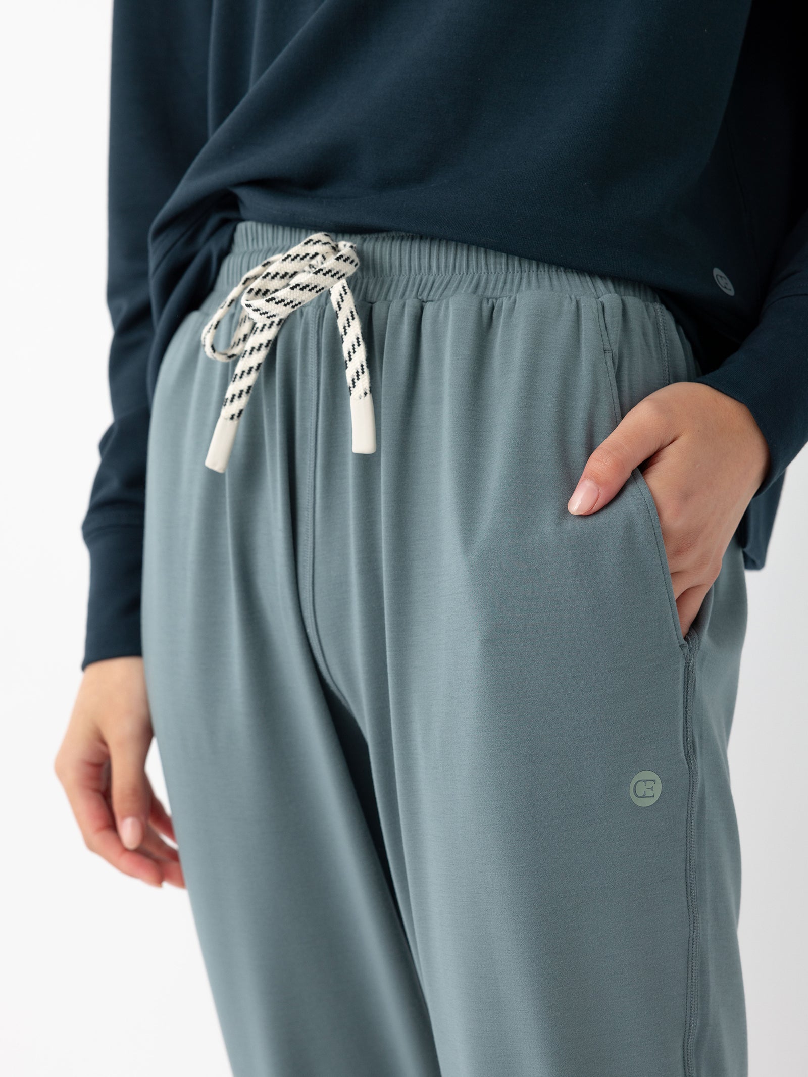 Close-up of a person wearing Cozy Earth's Women's Studio Jogger in light blue-gray with white drawstrings, paired with a dark blue long-sleeve top. The person's right hand is in the pocket, showcasing the joggers' casual and comfortable style.