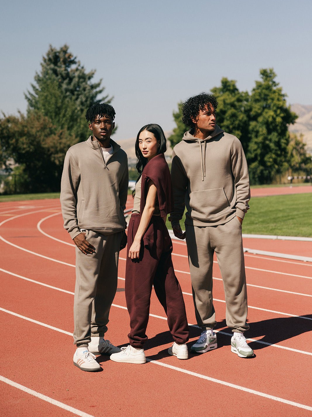 Three people stand on an outdoor track under a clear sky. Two individuals wear beige Men's CityScape Sweatpants from Cozy Earth, while the person in the middle wears maroon Men's CityScape Sweatpants from Cozy Earth. They all wear white sneakers and pose confidently with trees and a distant hill in the background.