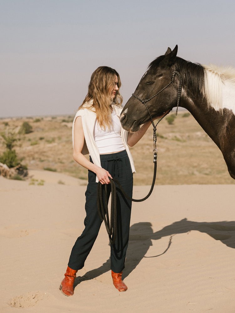 A woman in Cozy Earth's Women's Sunset Cropped Pant stands on sandy terrain, a sweater over her shoulders, holding the reins as a black and white horse nuzzles close.