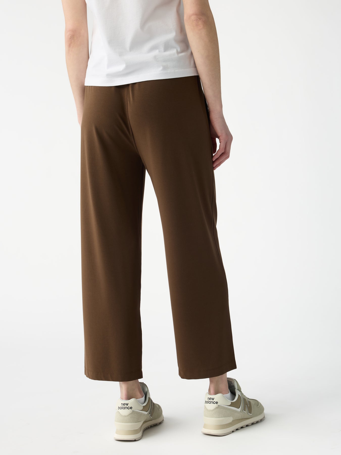 A person stands against a white background, wearing Cozy Earth's Women's Studio Cropped Wide Leg Pant in brown and white and gray sneakers, focusing on the pants and footwear from the lower half of the body.