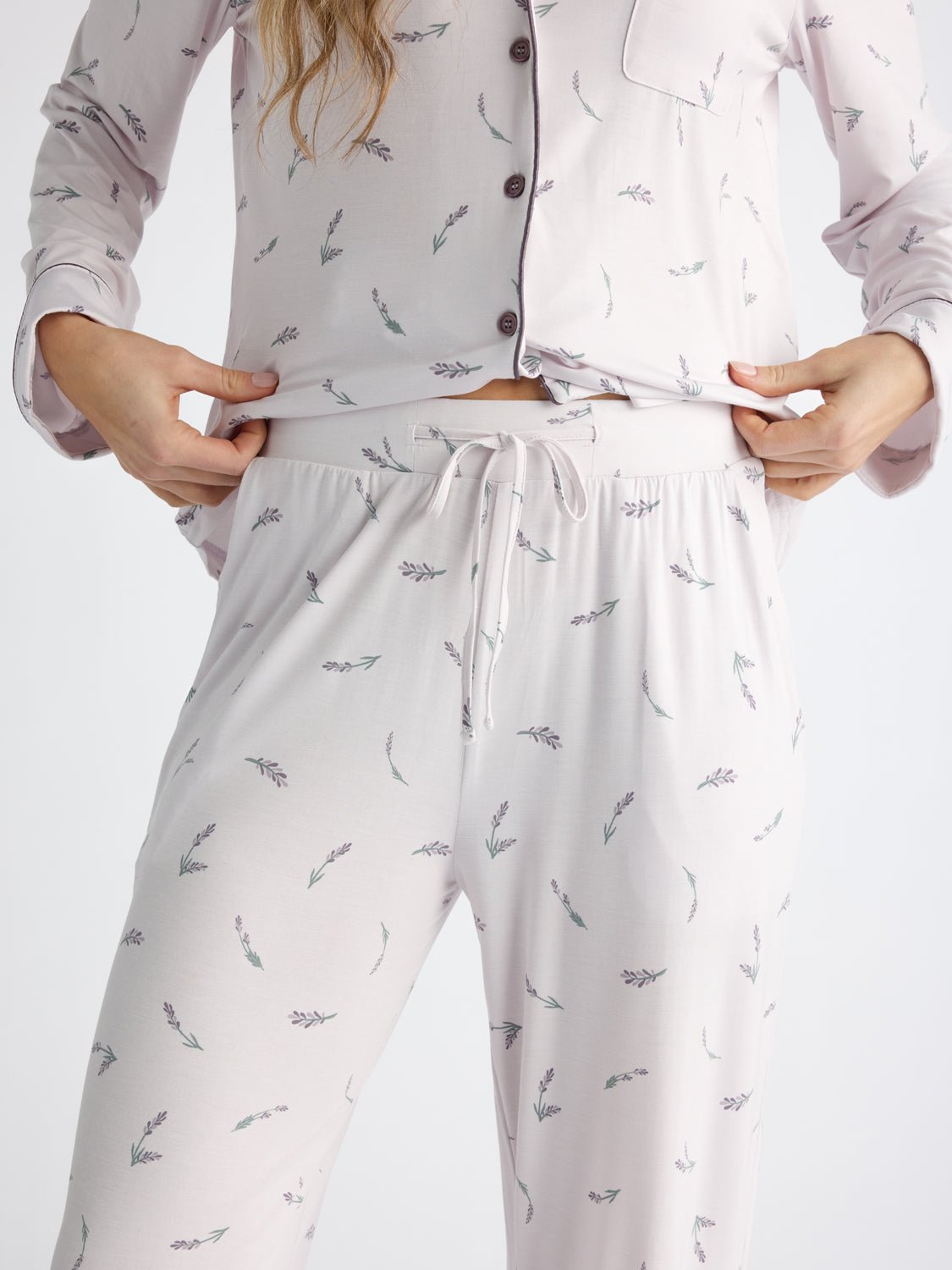 A person adjusts the waistband of Cozy Earth Women's Bamboo Stretch-Knit Pajama Pant, featuring a light color with small floral patterns. The top boasts buttons, a pocket, and both pieces offer a loose, comfortable fit.