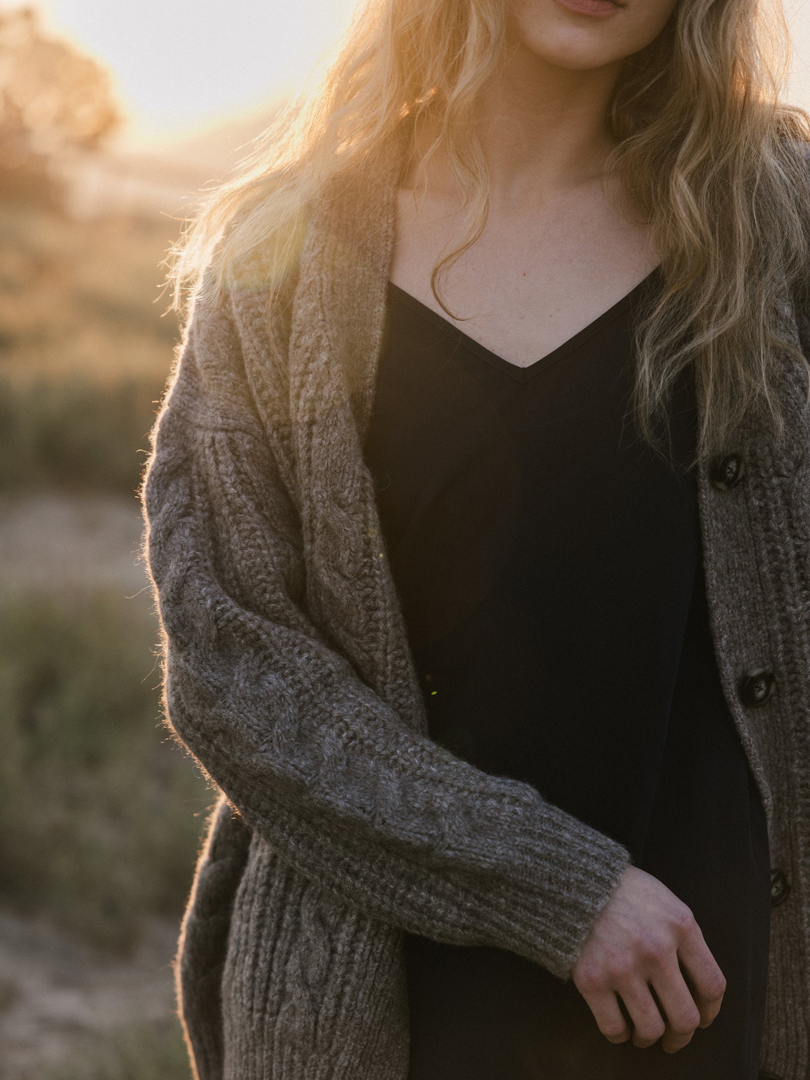 A woman with long blonde hair stands outdoors during golden hour, wearing a black dress and an Oversized Cable Knit Cardigan from Cozy Earth. The warm sunlight creates a soft glow, highlighting the natural setting with grass in the background.