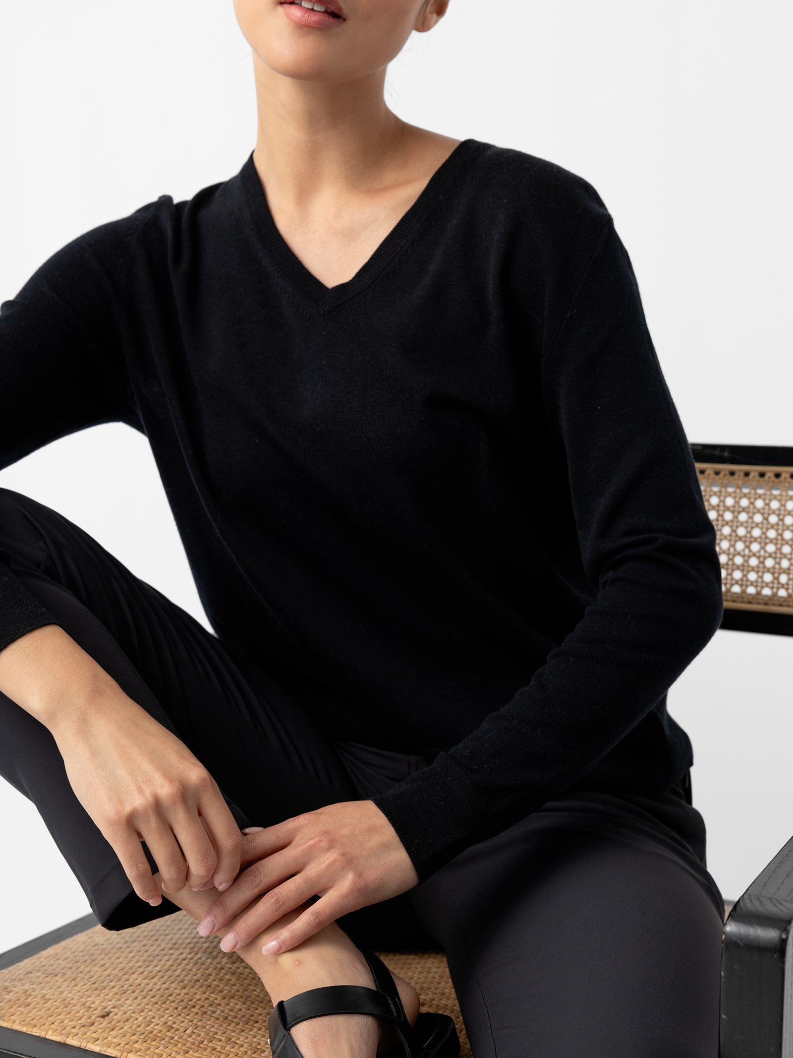 A person wearing a black Women's AirKnit V-Neck Sweater by Cozy Earth and black pants is seated on a woven cane chair. The individual has one arm resting on their knee, and the other hand placed on the chair. The background is plain white, and their face is partially out of frame.