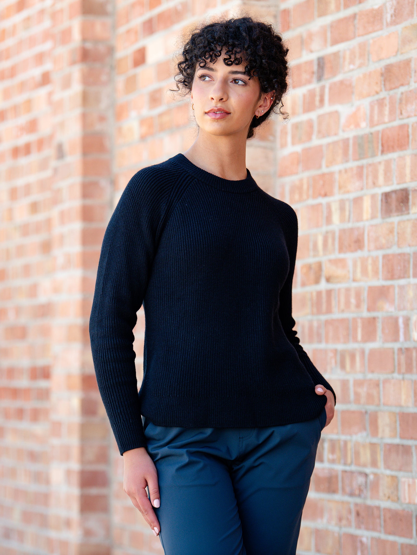 A person with curly hair is standing against a brick wall, looking to the side. They are wearing Cozy Earth's Women's Classic Crewneck in black and blue pants, with their hands in their pockets. The person has a contemplative expression.