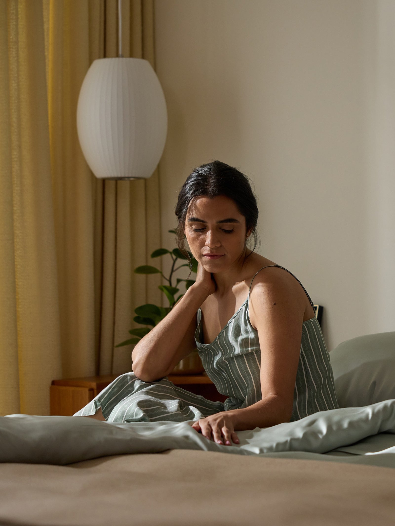 A woman sits thoughtfully on a bed adorned with a Cozy Earth Bamboo Duvet Cover in a softly lit room. Wearing a striped dress, she touches her neck gently. A lamp and small plant sit nearby against drawn curtains.