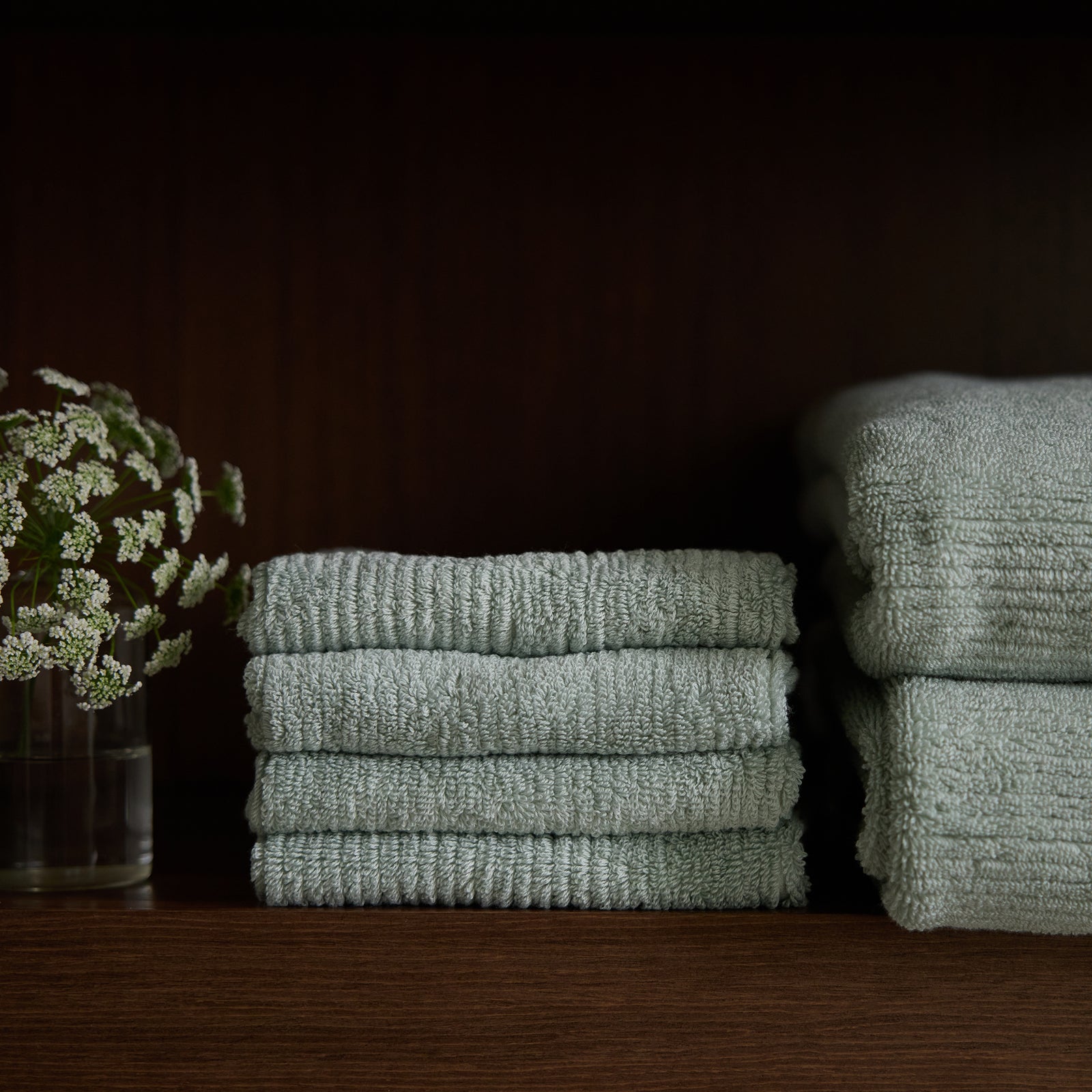 A neatly stacked pile of light green Ribbed Terry Washcloths by Cozy Earth sits on a dark wooden shelf. To the left of the washcloths is a clear glass vase holding sprigs of small white flowers. Another slightly out-of-focus stack of washcloths can be seen on the right side of the shelf.