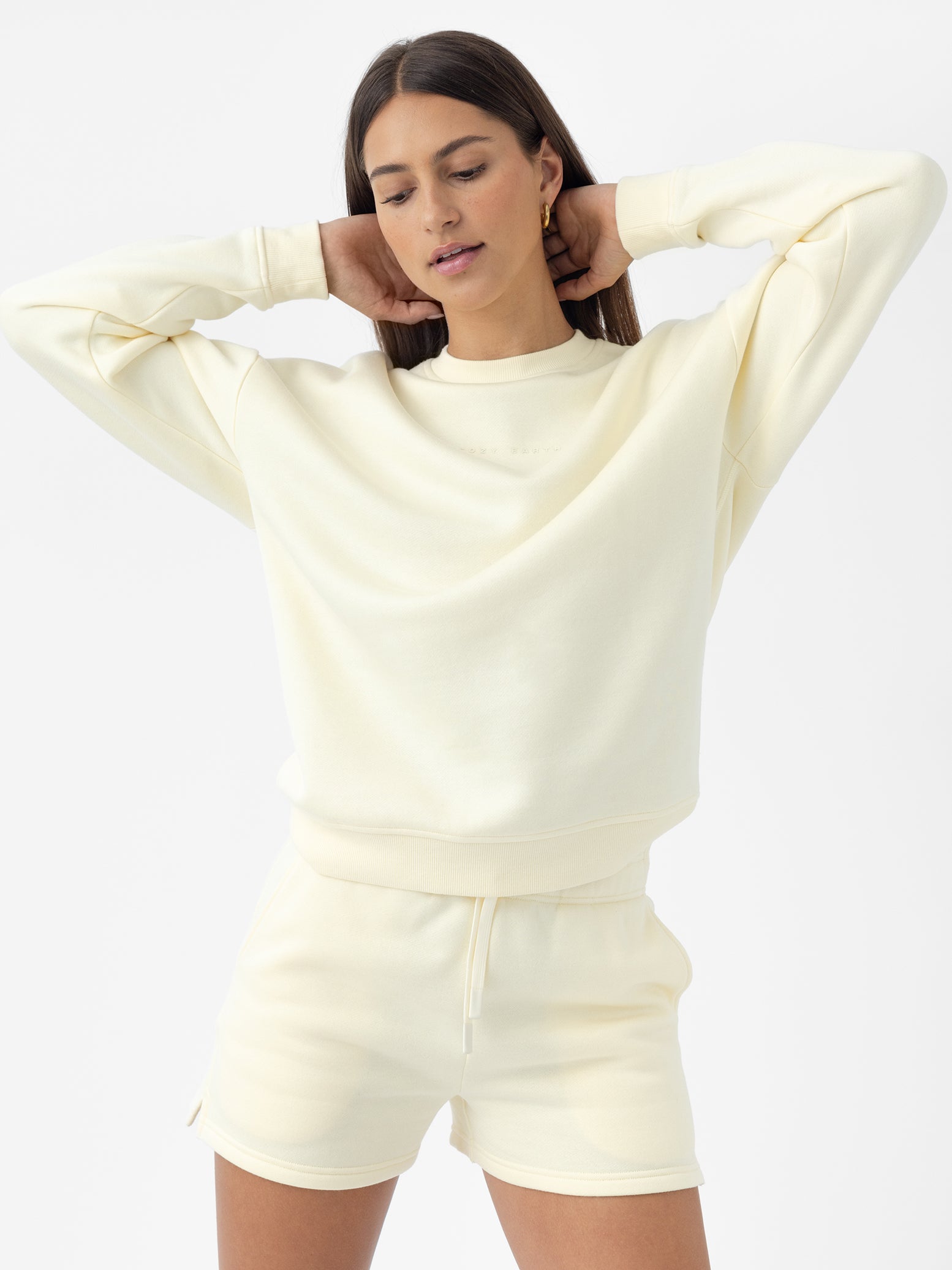 A person with long hair models the Women's CityScape Crewneck sweatshirt and matching shorts from Cozy Earth, posing with their hands behind their head against a plain white background.